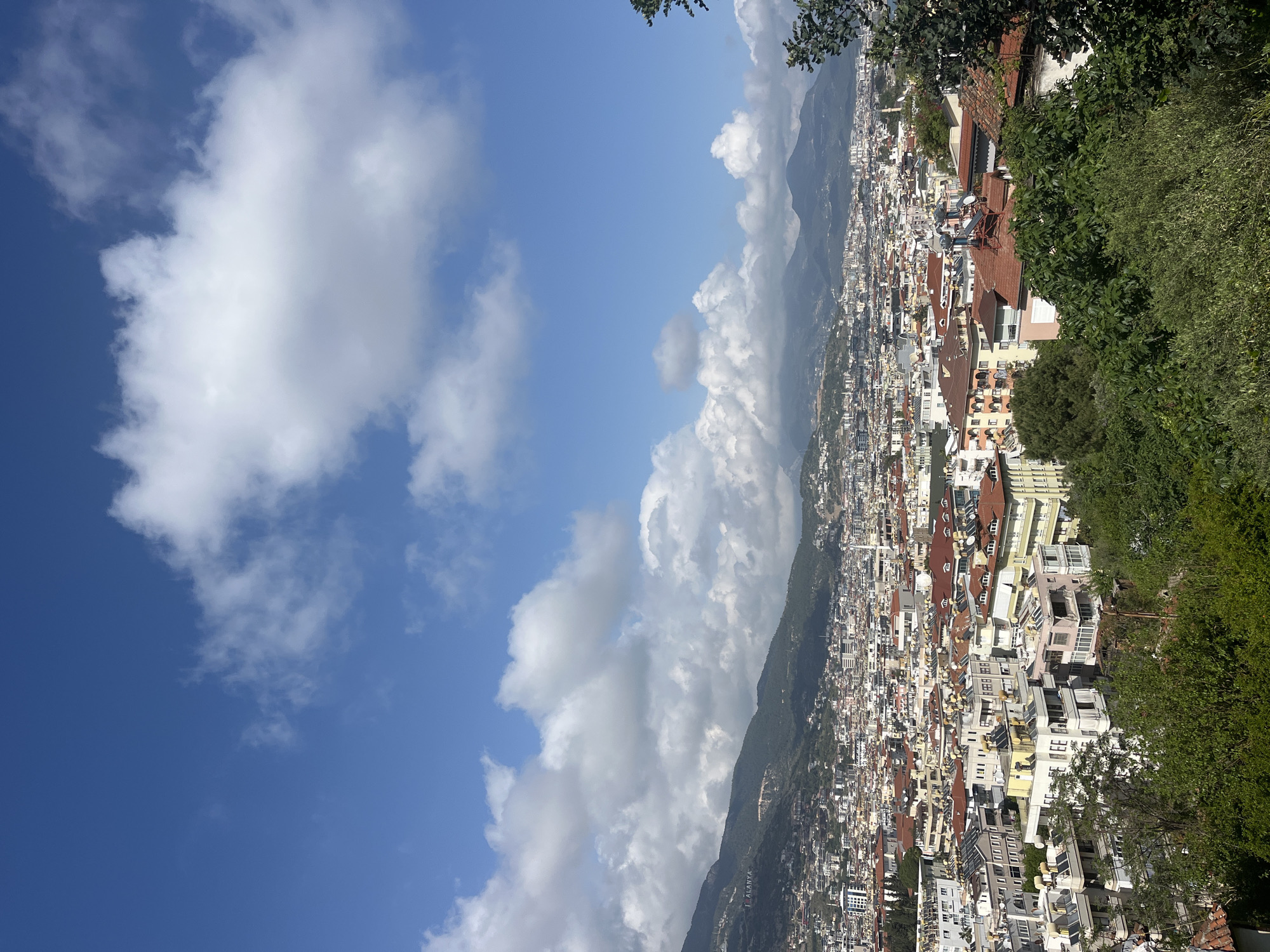 The image captures an elevated view of Alanya, Turkey. The foreground includes green foliage along the right side and top of the frame, suggesting the perspective is from a high vantage point. The city sprawls out below, characterized by dense urban development with buildings of varying heights and colors, including white, beige, and red-tiled roofs. The mountain ranges stand behind the city. The sky occupies the left half of the image and is a bright blue with large, fluffy white clouds.