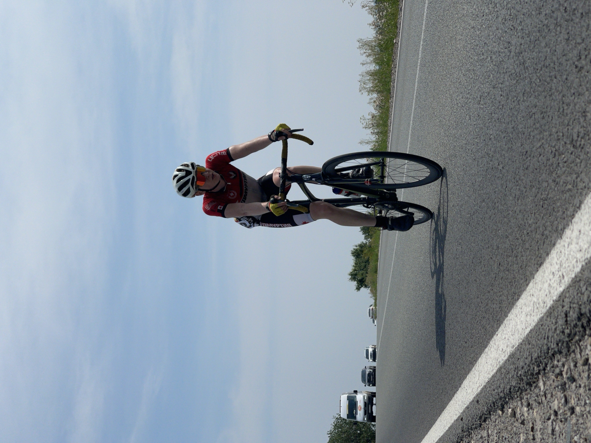 The image depicts a young male cyclist wearing a red and black cycling jersey, black cycling shorts, a white helmet, and orange sunglasses. He is actively cycling on an asphalt road under a bright, partly cloudy sky. The cyclist is positioned in the foreground, with his body angled slightly towards the viewer. In the background, a white van and several cars are visible on the same road. The environment appears to be rural, with grassy verges and vegetation lining the side of the road. The specific location in Perevoznoe, Russia, is not explicitly indicated by any visible landmarks or signage in the image. The overall scene captures a moment of athletic activity on a public road.