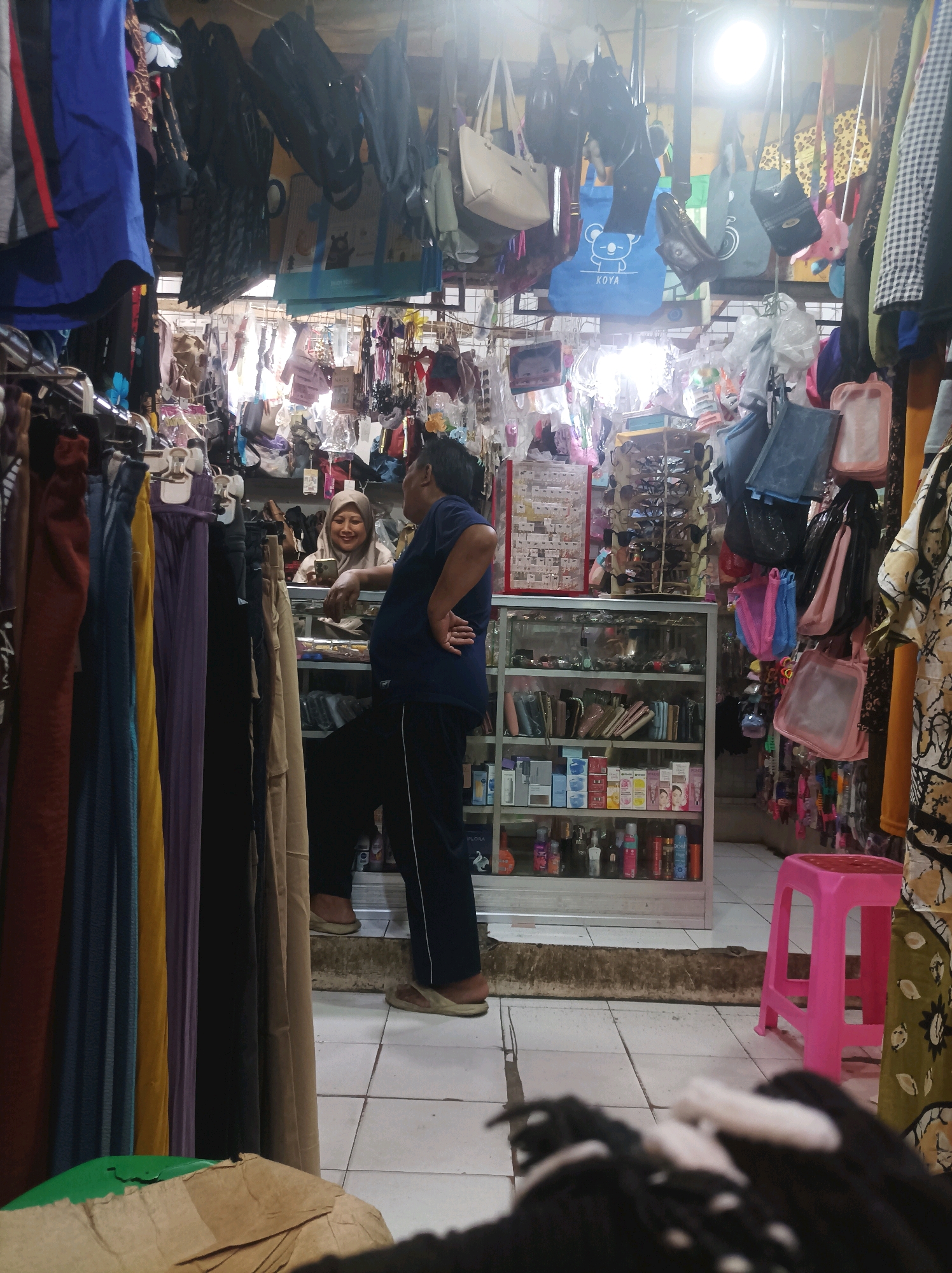 This image depicts a small shop interior in Sumuragung, Indonesia. The shop is densely packed with merchandise, with clothing and accessories hanging from the ceiling and displayed on shelves. In the foreground, colorful garments, possibly skirts or trousers, are hung on racks.
Two individuals are visible within the shop. On the left, a woman wearing a hijab is standing behind a counter, looking at a smartphone and smiling. She appears to be interacting with a man on the right, who is leaning against the counter with his hand on his hip, also looking towards the woman. The man is wearing a dark blue t-shirt and dark trousers.
Behind the counter, glass-fronted shelves display a variety of items, including cosmetics, perfumes, and small bags or wallets. Various handbags and other accessories are suspended from the ceiling throughout the shop. A bright pink plastic stool is positioned near the right side of the image. The overall environment suggests a busy, independent retail setting. The lighting is somewhat diffused, likely from overhead bulbs.