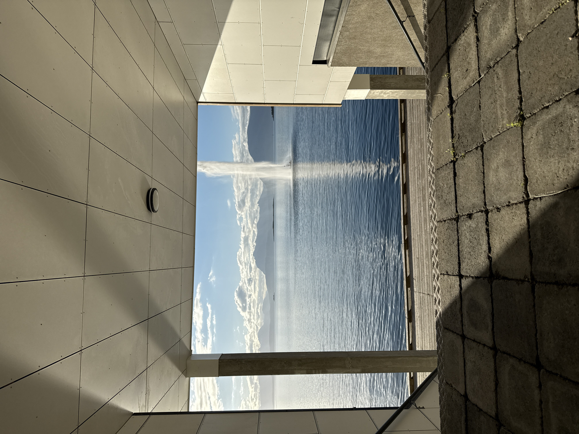 This image captures a scenic view of the harbor in Harstad, Norway, as seen from under a modern architectural overhang. The foreground is dominated by rough, gray paving stones with patches of green weeds growing between them, leading down a set of concrete stairs with a metal handrail. Beyond the stairs, a wooden boardwalk borders a tranquil body of blue water. In the distance, a tall water fountain sprays upwards, creating a dramatic vertical element against the horizon. The background features a range of majestic, silhouetted mountains under a bright blue sky dotted with fluffy white clouds. The overall scene is one of serene natural beauty juxtaposed with contemporary urban design, bathed in the warm light of what appears to be late afternoon or early morning.
