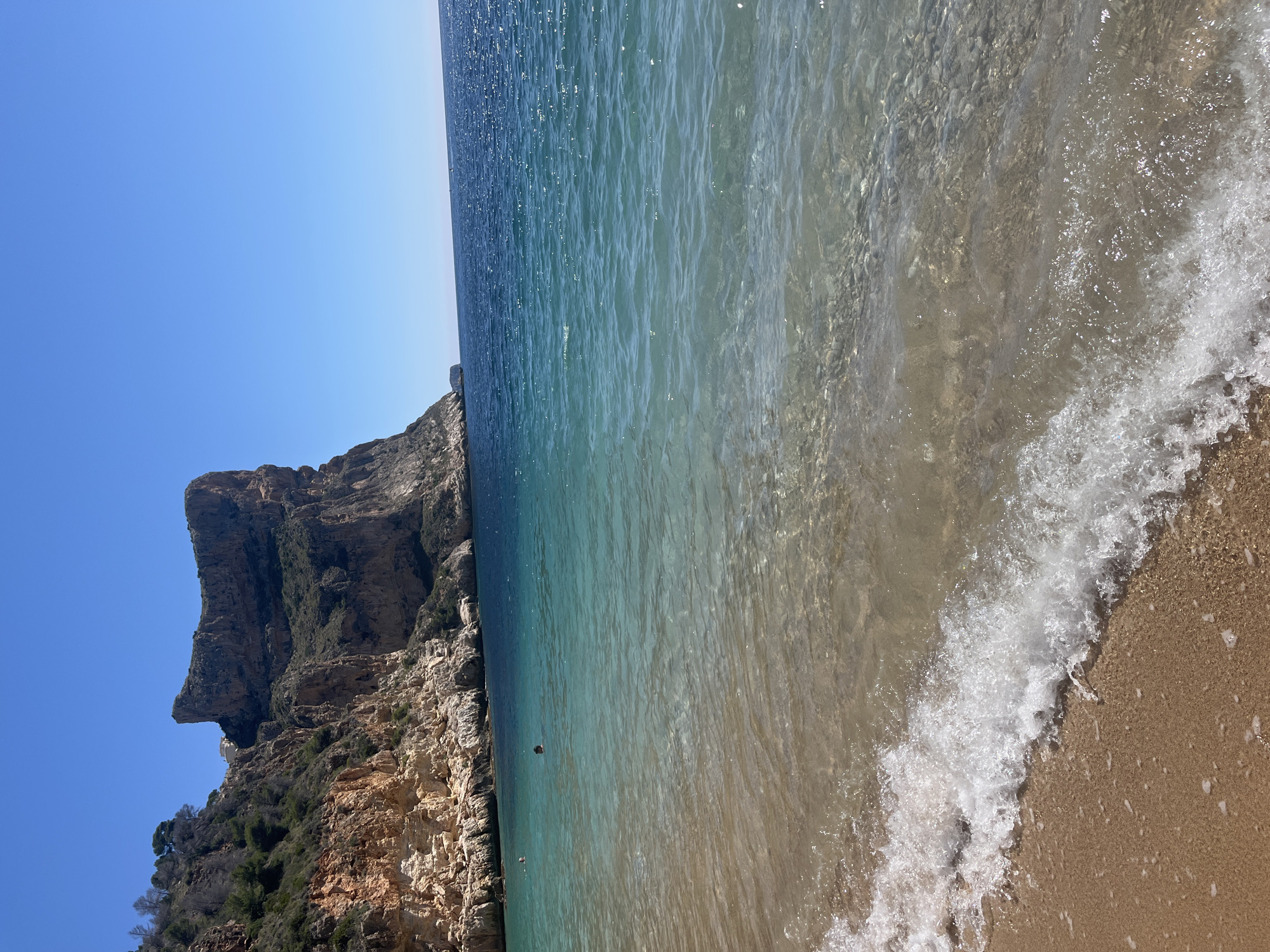 The image shows a coastal scene in el Poble Nou de Benitatxell / Benitachell, Spain. The foreground is dominated by a sandy beach and the clear, turquoise water of the Mediterranean Sea, with small waves lapping the shore. The water transitions from light near the shore to a deeper blue further out. To the left, a steep, rocky cliff rises sharply from the sea. The cliff is composed of layered sedimentary rock and partially covered with sparse vegetation. The sky is a clear, cloudless blue. The image is rotated counterclockwise.