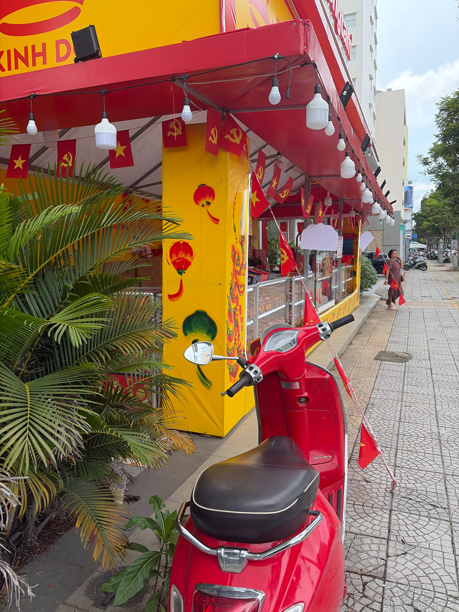 A red scooter is parked on a patterned sidewalk in front of a bright yellow building with a red awning. The awning is decorated with numerous small red flags featuring yellow stars and hammers and sickles, as well as red and yellow lanterns. Hanging from the awning are several lightbulbs, some exposed and some enclosed in white casing. A large palm tree is positioned to the left of the scooter, partially obscuring the building. Further down the sidewalk, a woman in a brown dress is walking away from the camera, holding a shopping bag and a red flag. Behind her, the street is lined with trees and buildings, and other scooters are visible in the distance, suggesting an urban environment in Da Nang, Vietnam. The sky is overcast.
