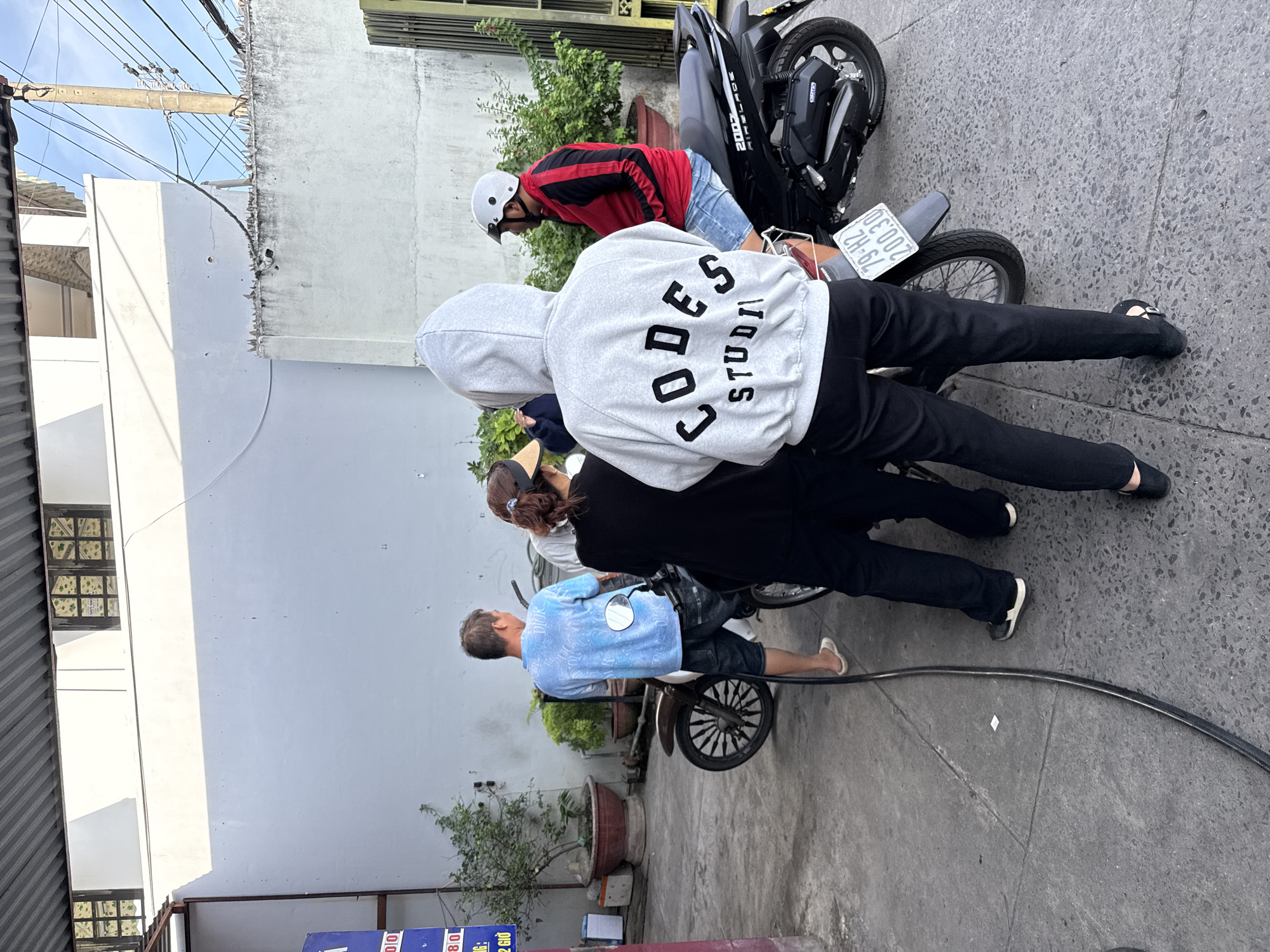 At a gas station in Bac Nha Trang Ward, Vietnam, three individuals are gathered around a motorcycle. The person in the foreground, facing away from the camera, wears a grey hooded sweatshirt with the text "CODES STUDIO" on the back and black pants. To their left, a man in a blue t-shirt and denim shorts stands near the motorcycle. A woman with a ponytail and a visor is partially visible behind the man. To the right of the foreground figure, another person sits on a separate motorcycle, wearing a red and black jacket, blue shorts, and a white helmet. A fuel hose is visible on the ground, suggesting refueling activities. The environment includes a concrete pavement, a white building with windows in the background, potted plants, and overhead roofing. The sky is visible in the upper right portion of the image.