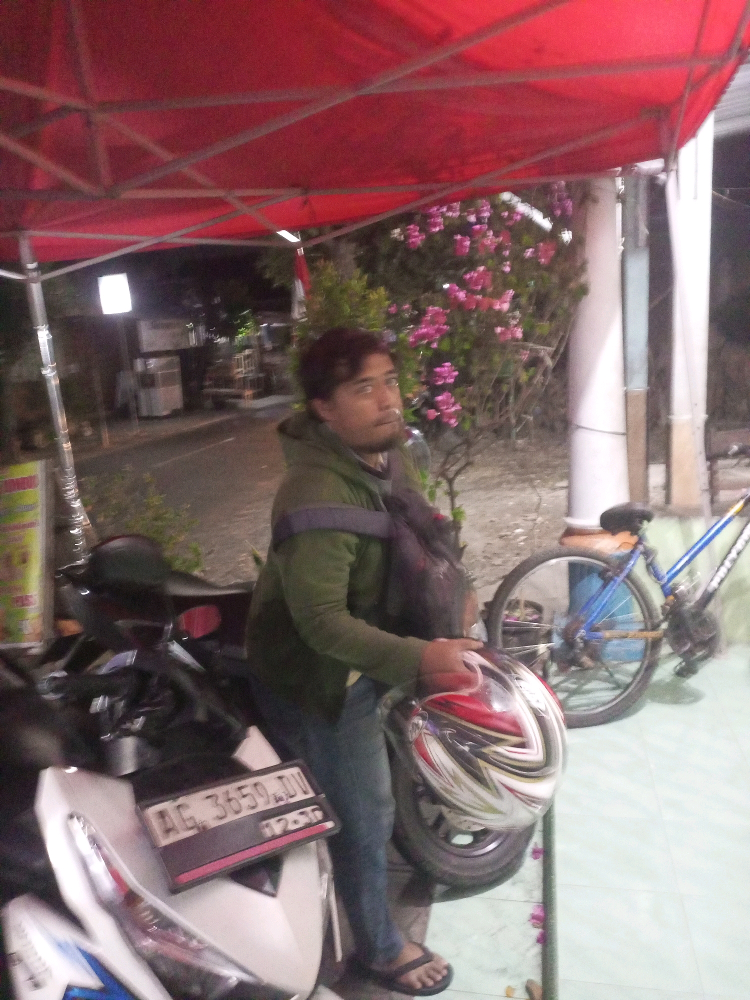 A man stands next to a motorcycle and bicycle under a red canopy. The man is wearing a green jacket and blue jeans. He is holding a helmet in his right hand. The motorcycle has a license plate that reads "AG. 3659. DD". The bicycle is blue and has a black seat. In the background, there are trees and buildings. The scene appears to be in a street in Kediri City, Indonesia, at night.