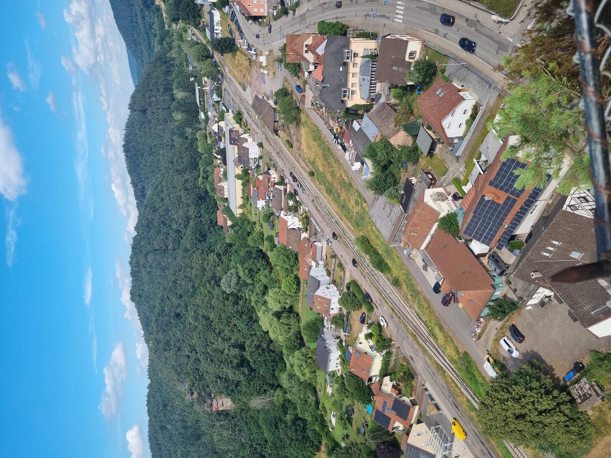 This is an aerial, long shot, daytime, eye-level, medium-wide, vertical, full-shot photograph of a residential area in Dahn, Germany. The scene is dominated by a dense, dark green forest that covers a steep hill on the left side of the image. A clear, bright blue sky with a few wispy white clouds is visible in the upper left quadrant.
To the right of the forest, a railway track runs diagonally across the frame. Adjacent to the railway, a road is visible with several cars parked along its edge and a few moving. A scattering of residential buildings, mostly with red-tiled roofs, are nestled between the forest, the railway, and the road. Some of these buildings have solar panels installed on their roofs. The houses appear to be of various sizes and architectural styles, with gardens and trees surrounding them. The overall setting suggests a semi-rural or suburban environment where nature and human habitation are in close proximity. There is no discernible activity or event taking place; the image captures a still moment. The weather appears to be fair and sunny, indicating a clear day. No text is visible in the image.