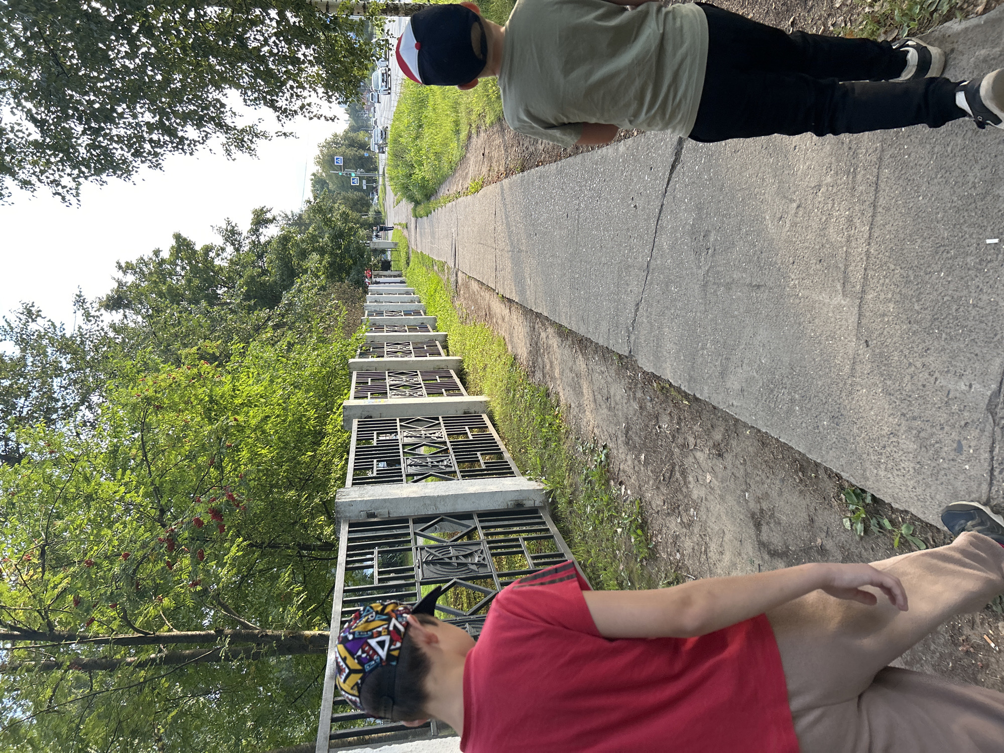 Two young boys are walking on a sidewalk in Chaikovsky, Russia. The boy on the left is wearing a red t-shirt, tan sweatpants, and a baseball cap with a colorful geometric pattern. He is walking away from the camera, facing a metal fence with a concrete base and decorative panels. Behind the fence, trees with green leaves and red berries are visible. The boy on the right is wearing a light green t-shirt, black pants, and a black and white baseball cap. He is walking away from the camera, also on the sidewalk. The sidewalk is made of concrete and runs alongside a grassy area and the fence. In the background, a road with cars and a pedestrian crossing signal can be seen, indicating an urban environment. The sky is bright, suggesting daytime.
