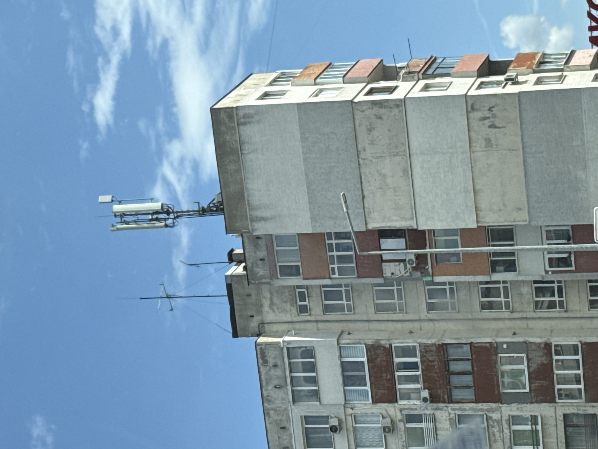 A rotated, medium-angle, eye-level shot depicts a tall, dilapidated residential building in Sofia, Bulgaria, set against a backdrop of a partly cloudy blue sky. The building, constructed in a Soviet-era style, features a mix of concrete panels and numerous windows, some with visible air conditioning units. Many of the window frames are painted brown. An array of communication antennas and a street light are attached to the top and side of the building, suggesting its use for telecommunications infrastructure. The building's weathered facade indicates age and potential lack of maintenance.