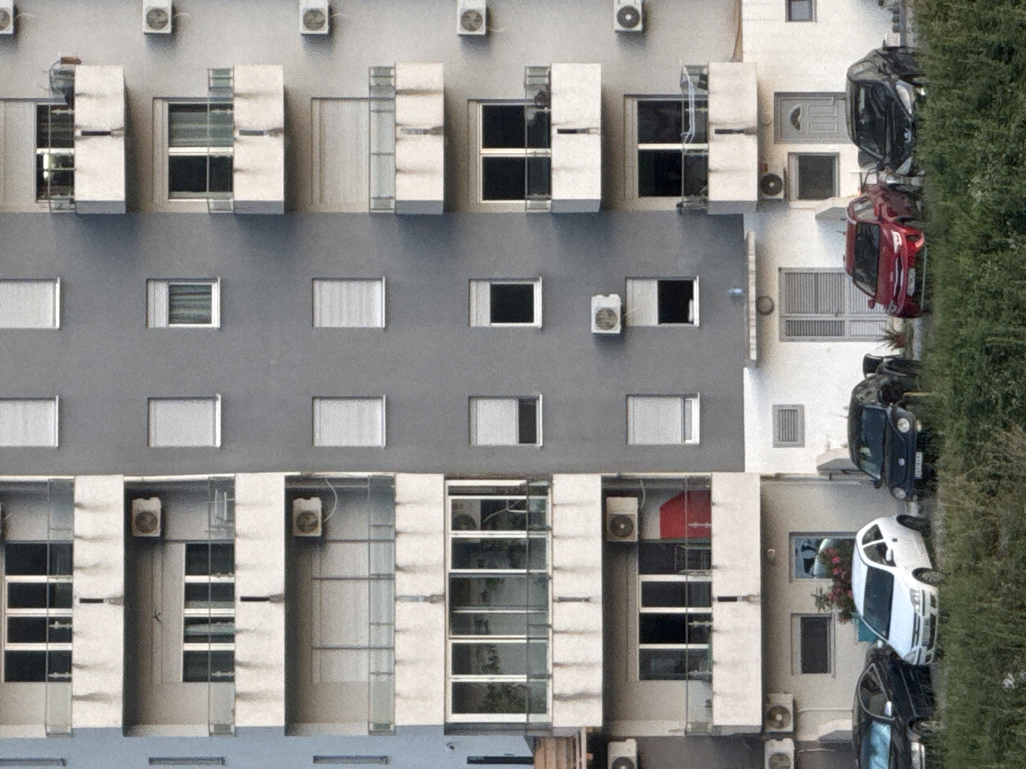 The image displays the exterior of a multi-story apartment building in Rafailovići, Montenegro. The building has a gray facade with multiple balconies and windows. Several air conditioning units are mounted on the exterior walls. At the ground level, there are three cars parked on a patch of grass. A black sedan is on the left, a white hatchback is in the center, and a black compact car is on the right. The cars are parked in front of the building's ground floor entrance and windows. The surrounding area is overgrown with green grass and vegetation.