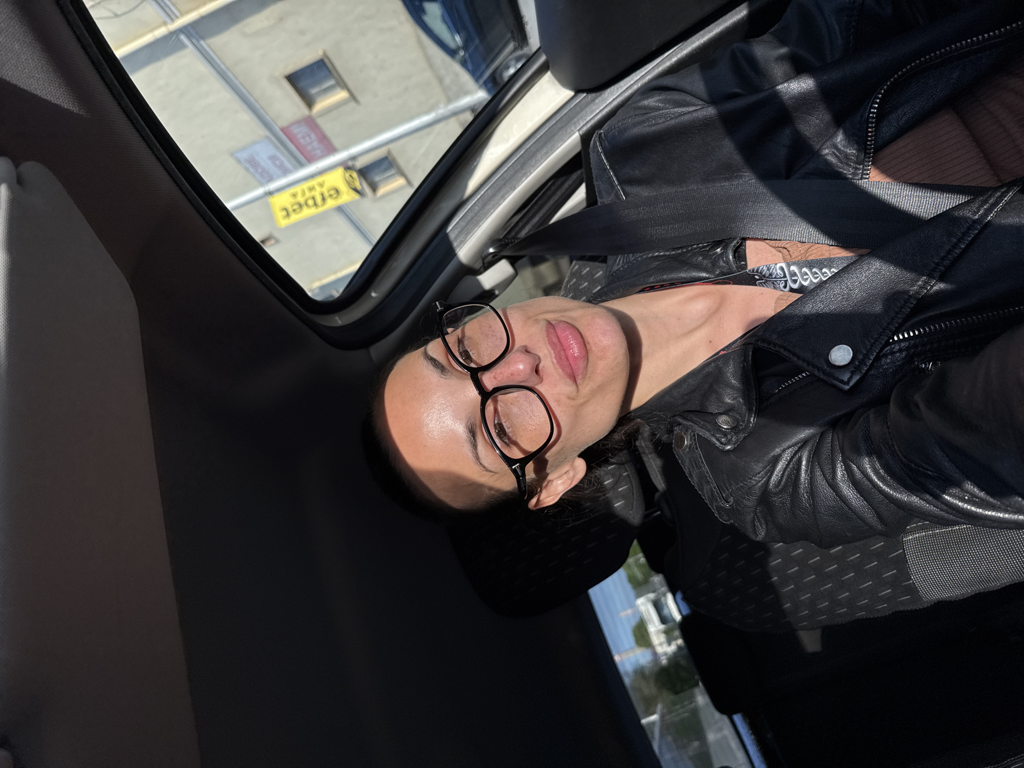 The image, captured from a low angle, presents a woman seated in what appears to be the front passenger seat of a car. Her head, adorned with dark-rimmed glasses, is positioned on the right side of the frame, looking upwards and slightly left. She wears a black leather jacket, with a safety belt crossing her chest. Strong, direct sunlight illuminates her face, creating prominent highlights on her skin and casting sharp shadows. Her dark hair is pulled back, and her expression is neutral with slightly parted lips.
Through the car's windshield or sunroof, a bright exterior scene is visible above and to the left of the woman. This includes parts of a building and a distinctive yellow advertising sign. The text on this sign is seen inverted and possibly mirrored. Clearly discernible Cyrillic letters spell out "АКЦИЯ" (AKCIA), which translates to 'Sale' or 'Action', alongside another word "erbet". The intense sunlight suggests a clear, sunny day, likely during the morning or afternoon. The presence of Cyrillic text on the sign acts as a key location cue, consistent with the provided context of Pleven, Bulgaria. The overall scene suggests a moment captured candidly or a selfie taken while traveling in a vehicle during daylight hours.