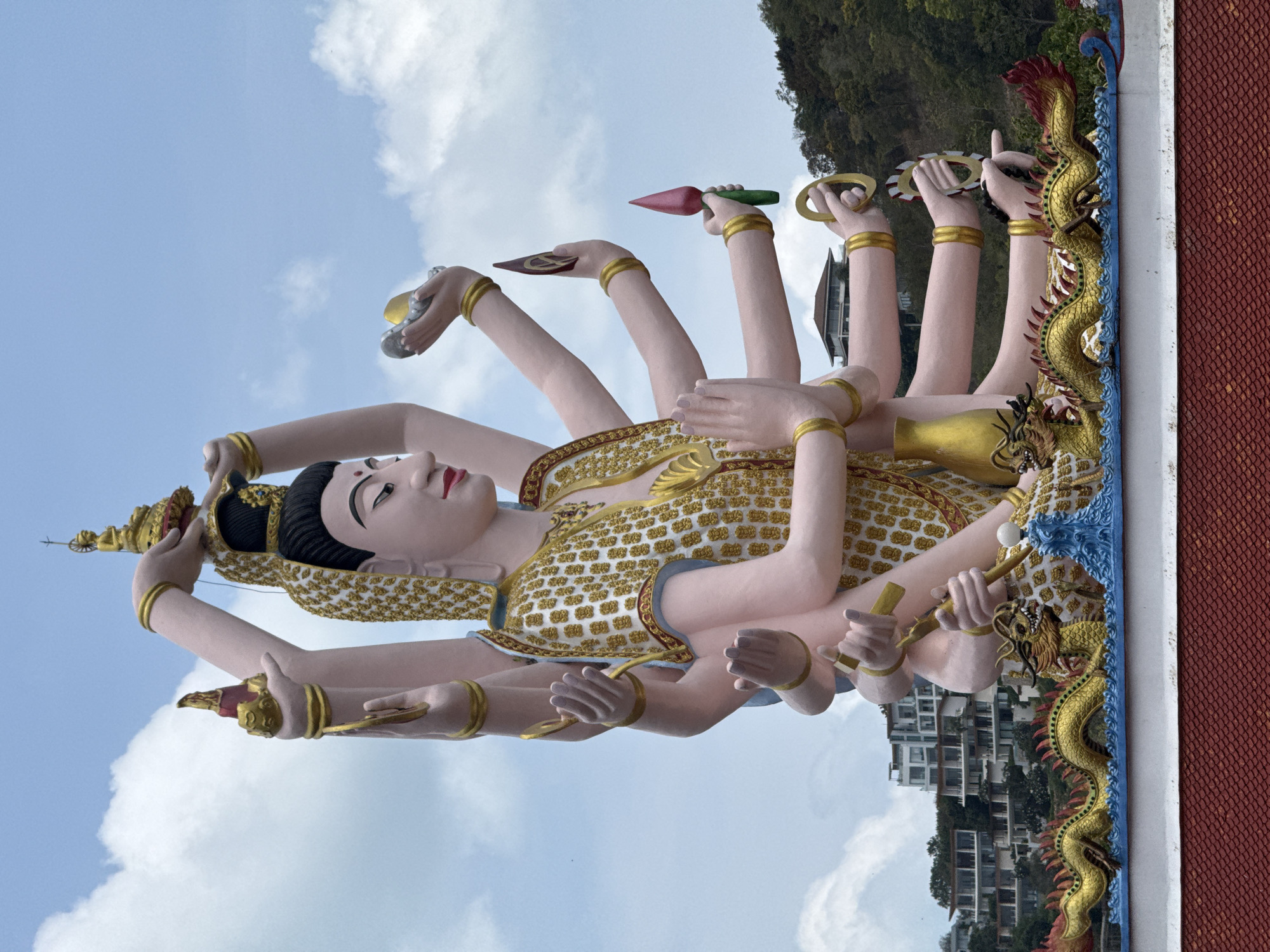 A large, multi-armed statue of a deity stands against a blue sky with scattered clouds. The statue is pink with gold accents, wearing an ornate gold garment. It has many arms, some of which are holding objects like a lotus flower, rings, and a bowl. The statue is positioned in front of a hillside with lush green trees and modern buildings. Below the statue, there is a decorative ledge featuring a golden dragon sculpture and a blue wave-like design. The foreground shows a red tiled roof. The overall scene suggests a religious or cultural landmark in Ko Samui District, Thailand.