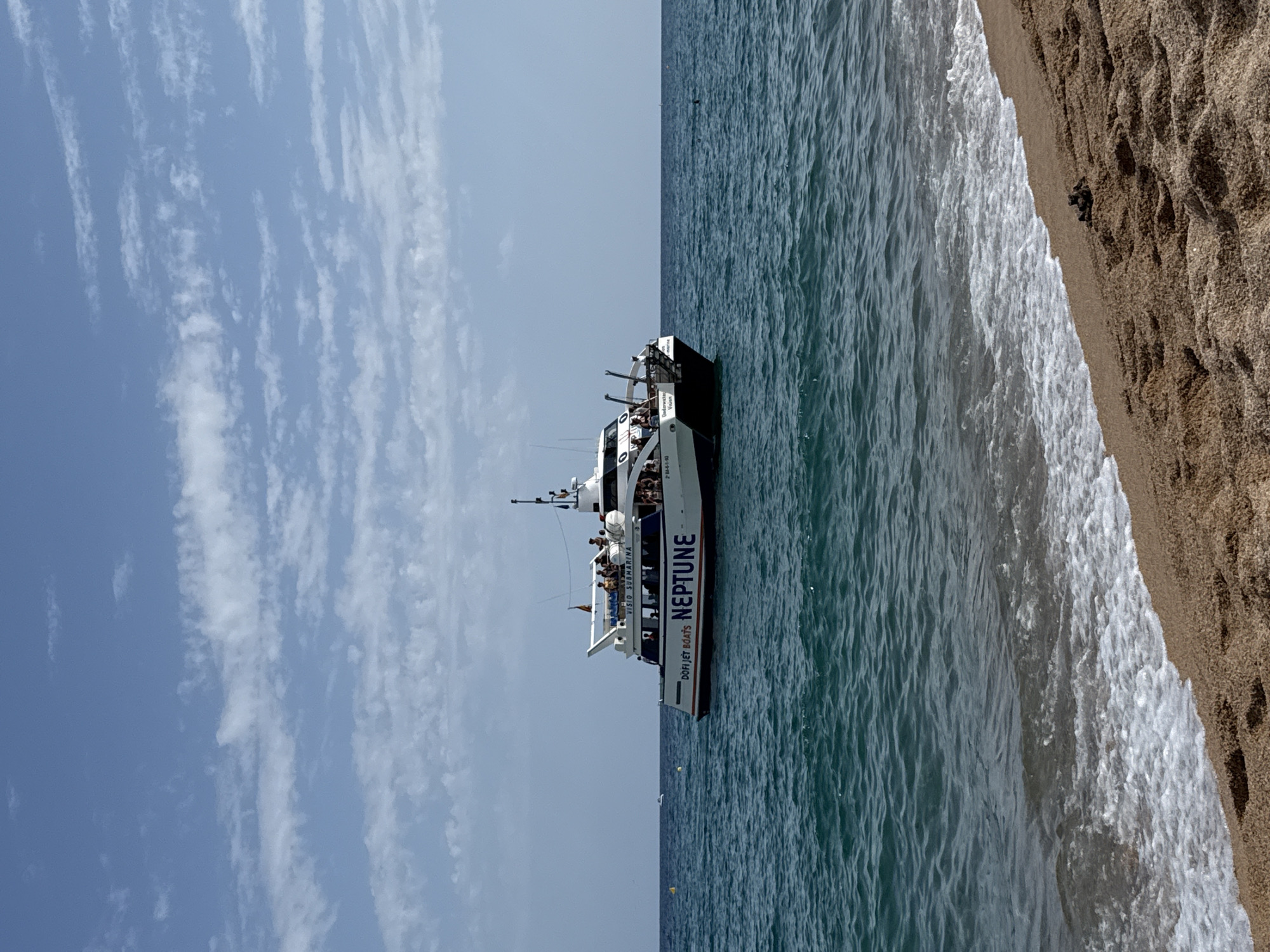 A white passenger boat, labeled "NEPTUNE" in large purple letters on its side, cruises on the blue sea near the shore of Blanes, Spain. The boat, which appears to be a ferry or excursion vessel, has multiple decks and is populated with passengers. The foreground shows a sandy beach with gentle waves lapping at the shore. The sky above is a clear blue with scattered white clouds. Two yellow buoys are visible in the water to the left of the boat, and a small white object, possibly another boat, is further out at sea on the left horizon. The overall scene depicts a sunny day at the coast.