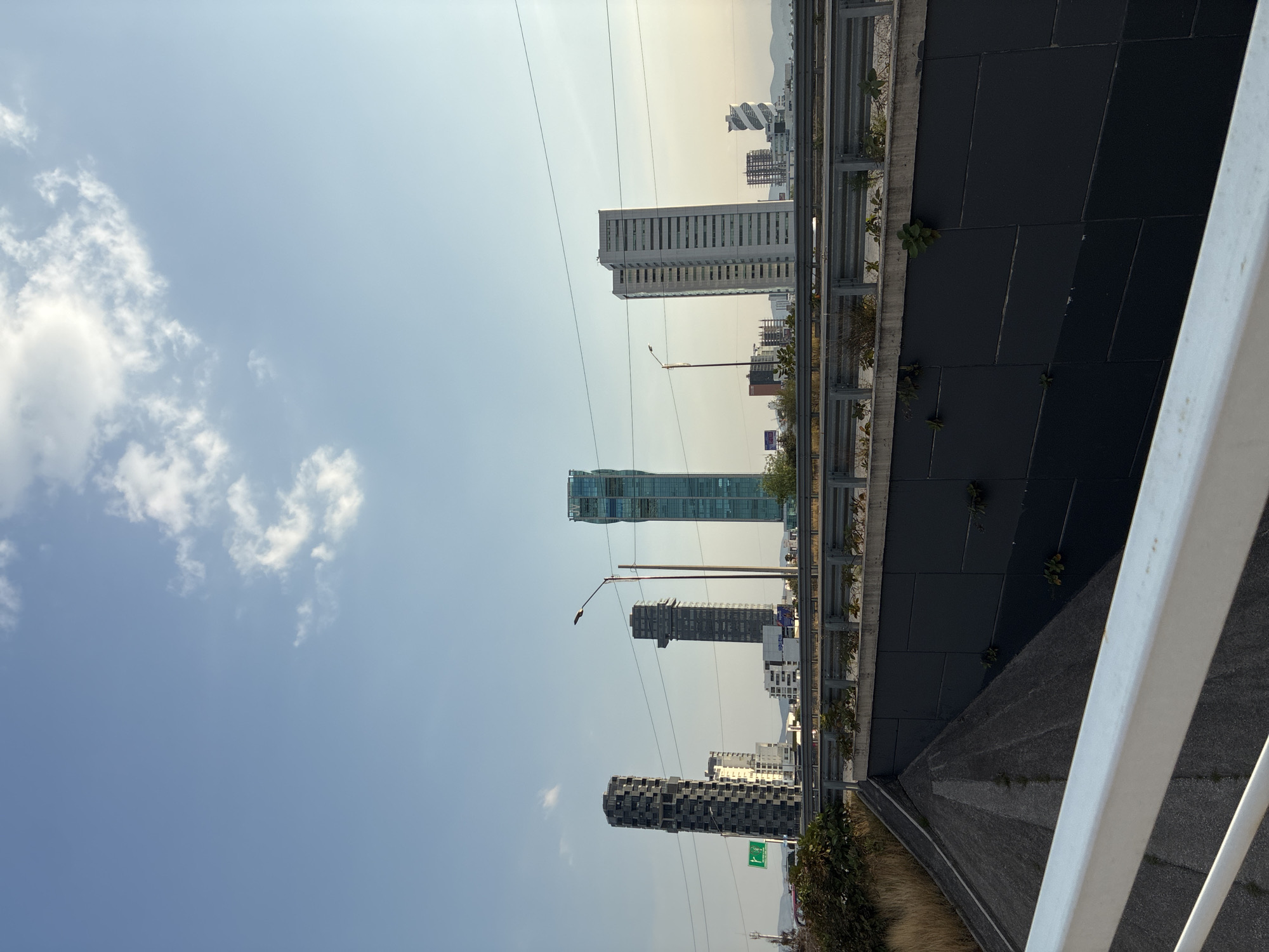 The image shows a view from an elevated position, likely a bridge or overpass, overlooking a cityscape. Several tall buildings are visible in the distance. The sky is light blue with scattered clouds. A highway barrier is in the foreground, constructed of gray metal. Some greenery is growing along the edge of the structure. The location is identified as San Bernardino Tlaxcalancingo, Mexico.