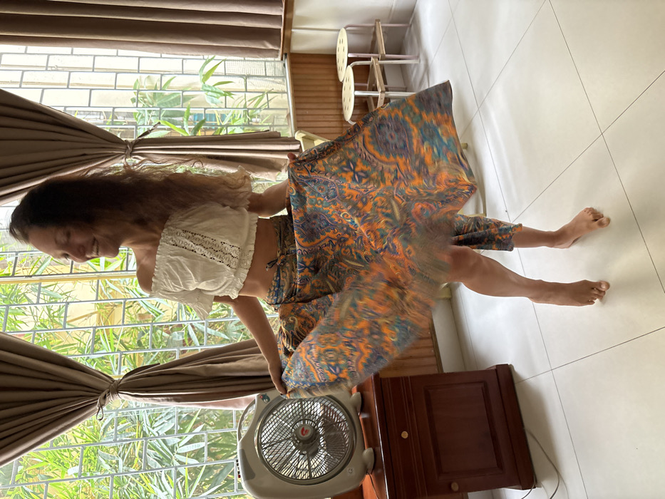 An adult female is depicted mid-motion within an indoor space, consistent with a residential interior in Hà Nội, Vietnam. The woman, with fair skin and long wavy brown hair, wears a white off-the-shoulder crop top featuring lace detail and a flowing, barefoot, multi-patterned skirt in orange, blue, and purple, blurred due to her spinning action. She smiles broadly with closed eyes and her head tilted back, indicative of enjoyment. Her left foot is flat, while her right foot is poised on its toes. To her left, a white box fan with a circular grille rests on a dark brown wooden nightstand with two drawers and gold-colored knobs. The floor is composed of white square tiles. The background features a large window with a metal security grille, framed by tied-back tan curtains. Visible through the window are dense green foliage and architectural elements of another building, suggesting an urban environment. Light brown wooden paneling extends below the window. Two light-colored stools with metal legs are visible in the background to the right. The scene is brightly illuminated by natural light entering through the window.