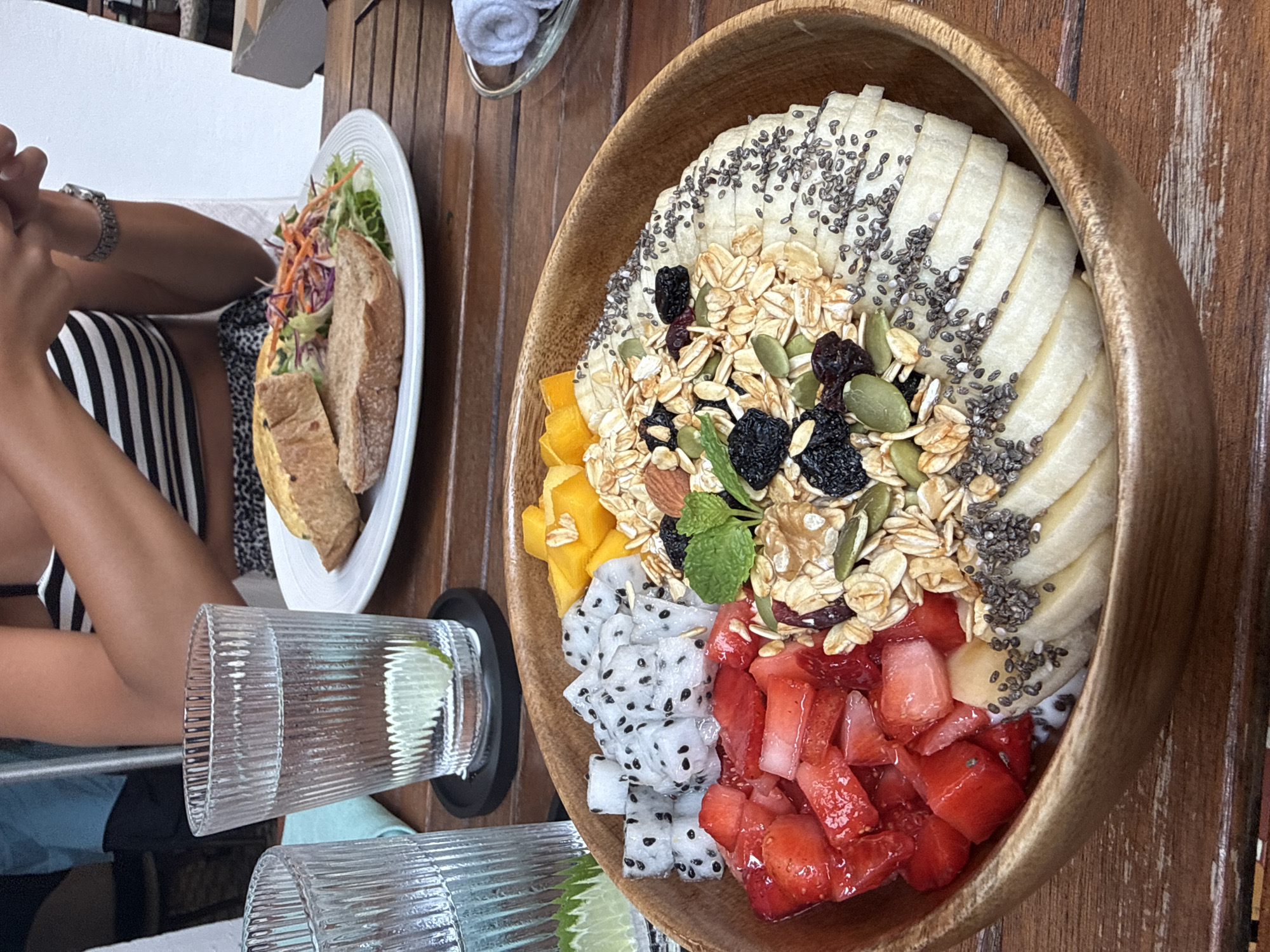 The image depicts a meal at a cafe, likely in Đà Nẵng, Vietnam given the prompt.
**Subjects and People:**
- A person is visible on the left, wearing a black and white striped garment.
- The person is sitting at the table and appears to be using a phone.
**Objects:**
- A large, wooden bowl filled with a fruit salad. The salad consists of sliced bananas covered with chia seeds, granola, mango, dragon fruit, and diced strawberries. The salad ingredients are arranged in sections within the bowl.
- A white plate with a sandwich and salad is placed nearby.
- Two glasses with clear liquid and lime wedges are in front of the bowl.
- A folded white towel is present in the background.
**Environment:**
- The setting is a cafe or restaurant, as suggested by the presence of food, drinks, and table service.
- The table surface is made of wood.
- The lighting is natural, likely from an outdoor source.
**Actions and Interactions:**
- The person is seated at the table and interacts with their phone.
- The food is arranged on the table, indicating a meal is either in progress or prepared for consumption.