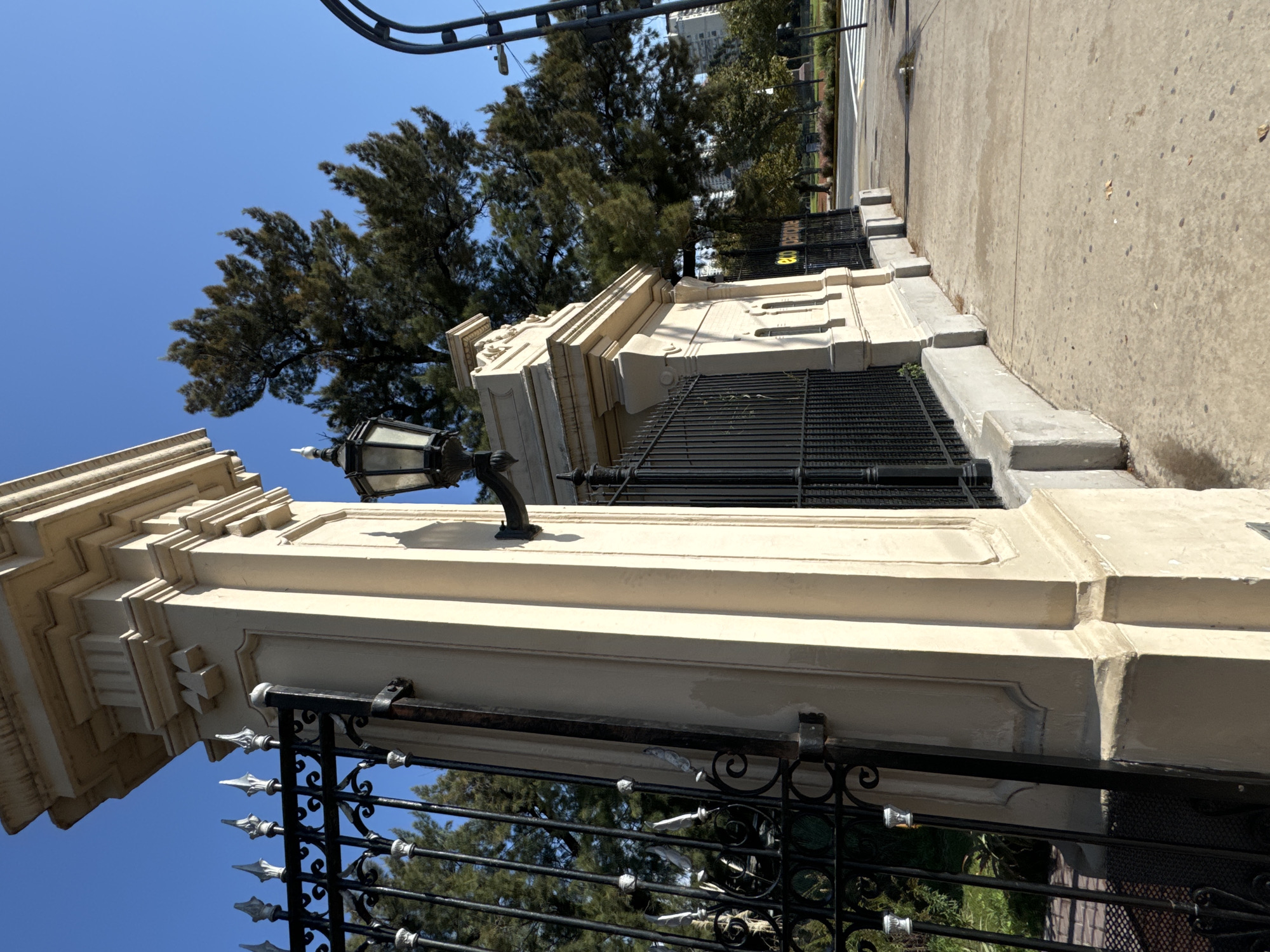 The image shows a portion of a cream-colored, ornate stone gate and fence with black ironwork in Buenos Aires, Argentina. The gate is constructed with decorative moldings and a stone lamp fixture. The fence features spear-tipped vertical bars and decorative wrought iron elements. Beyond the gate, trees are visible against a blue sky. A sidewalk and street are also visible. The picture is taken at an angle, looking upwards.