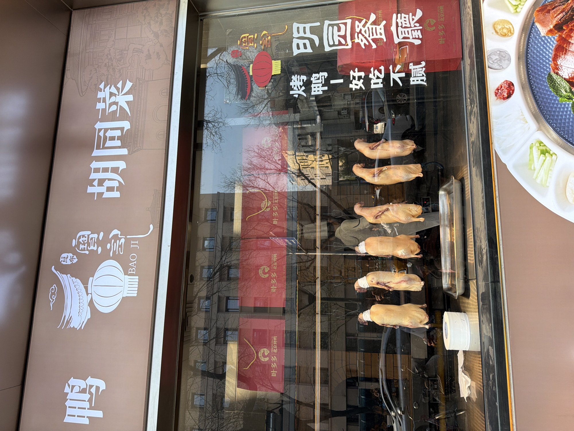 The image shows the exterior of a restaurant in Dongcheng District, China. The restaurant is called "Bao Ji", as indicated by the sign on the left. The sign also displays the phrase "胡同菜", which translates to "hutong cuisine". The window of the restaurant showcases Peking ducks hanging on hooks, ready to be roasted. The ducks have bandages around their necks. Another sign on the window indicates that the restaurant also serves "Ming Yuan Restaurant" cuisine. The reflection in the window reveals a cityscape with apartment buildings and bare trees. A sign for "King Duck" and "Ye Duoduo" (叶多多) is also visible, suggesting that the restaurant might specialize in Peking duck. Part of a plate of food that features sliced peking duck is also visible. The overall scene presents a typical Chinese restaurant storefront specializing in Peking duck dishes in the Dongcheng District.