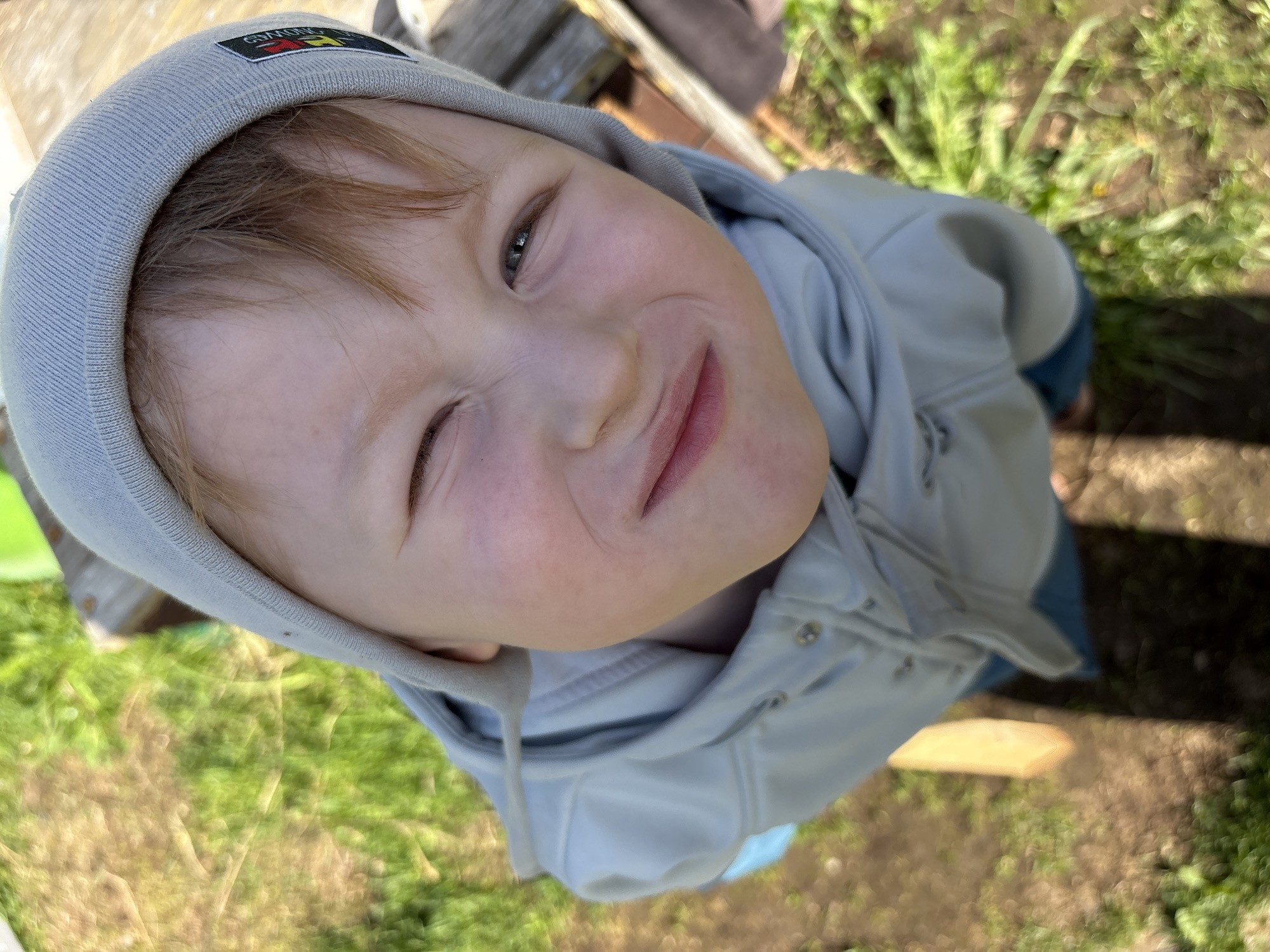 A young child with light skin and reddish-blond hair is wearing a light grey hooded jacket and matching beanie with a colorful patch. The child is looking up at the camera with one eye open and the other squinted shut. The photo is taken outdoors in an area with green grass and some visible wood. The location, Italmas, Russia, is likely relevant to the ethnicity/origin of the child.