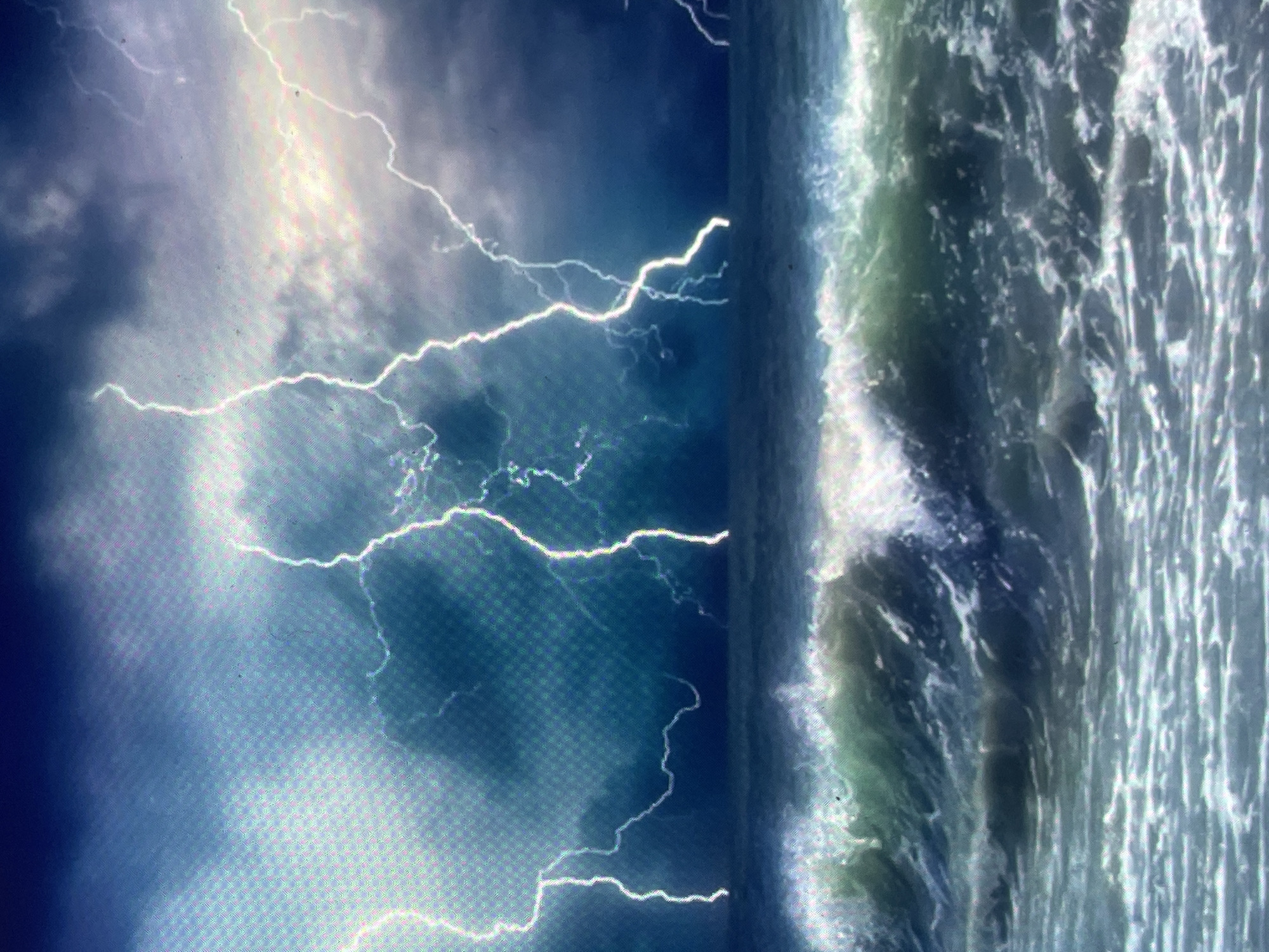 The image presents a turbulent oceanic storm scene, digitally captured with a noticeable grid-like pattern characteristic of a screen display. The upper portion of the frame depicts a dark, heavily clouded sky, exhibiting deep blue and grey tones consistent with severe weather. Numerous bright white and light blue lightning bolts descend from the stormy heavens towards the tumultuous ocean surface below. The middle and lower sections of the image are dominated by rough sea conditions, featuring large, breaking waves with powerful white caps and foamy crests. The water itself varies in color from dark green-blue in the troughs to frothy white at the peaks. No human presence, marine vessels, or landmasses are discernible. The specified location for this scene is Zelenyi Hai, Ukraine.