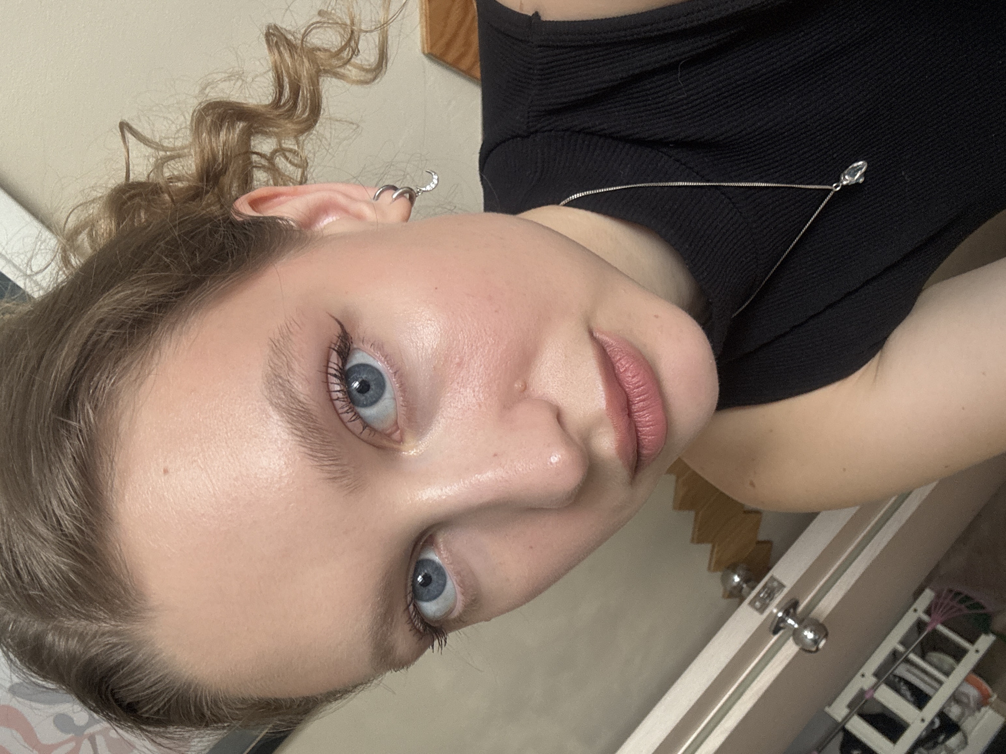 The image shows a close-up of a young Caucasian woman's face. She has blue eyes, light skin with some visible pores and a few small blemishes. Her eyebrows are light brown and well-defined, and she is wearing dark eyeliner with a slight winged tip. Her lips are slightly parted and appear to be a soft pink color, with subtle lip liner. Her blonde hair is styled in loose curls, pulled back from her face. She is wearing a black sleeveless top, a silver necklace with a small pendant, and a silver hoop earring in her left ear with a small crescent moon charm. The background is out of focus but appears to be an indoor setting with a light-colored wall and some wooden elements. There are no discernible landmarks or specific details that would allow for a precise location to be identified within the city of Izhevsk, Russia.