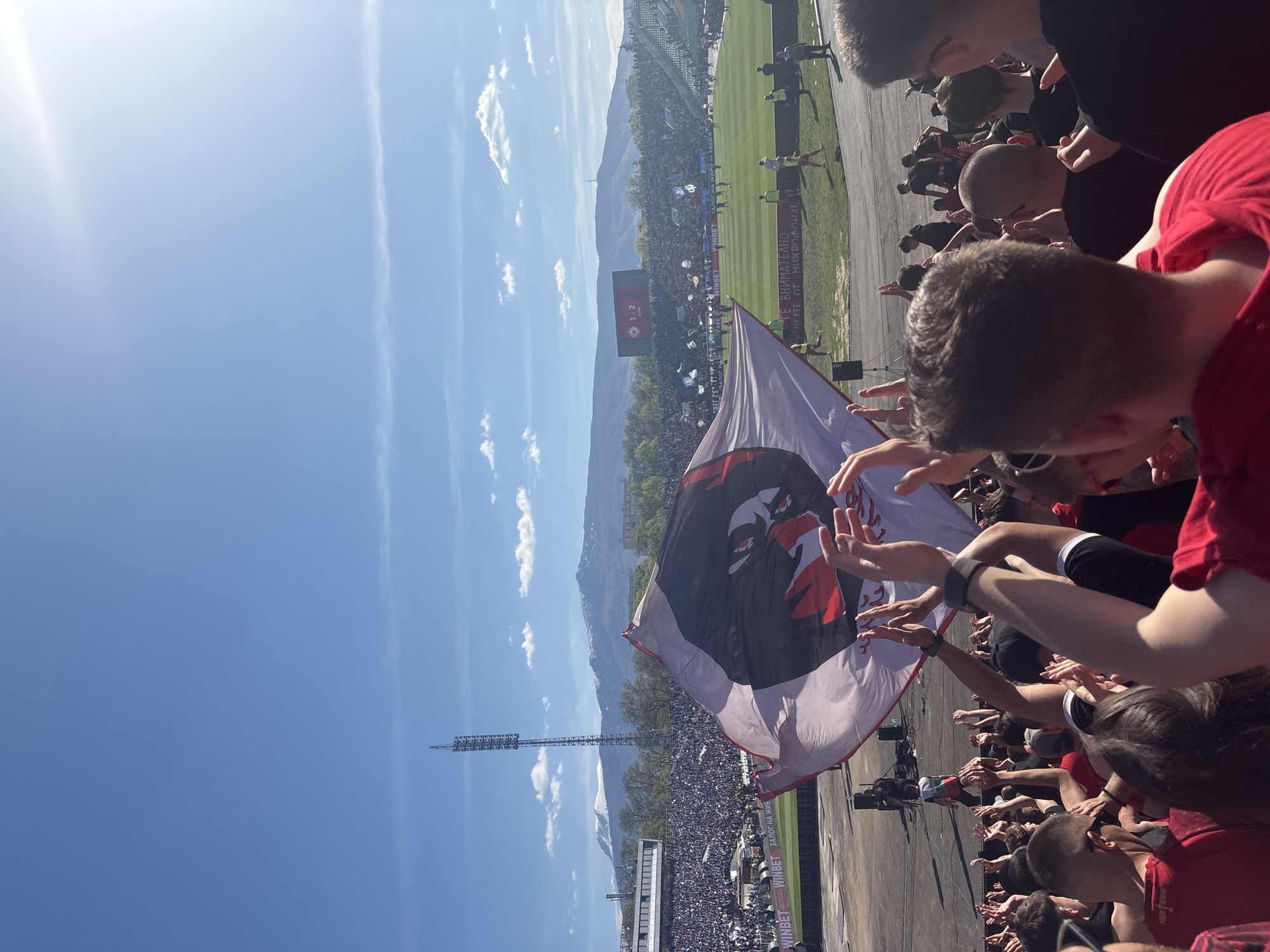 The image shows a crowd of people, likely at a sporting event, holding up a large flag with a black and red design. The event appears to be outdoors on a sunny day. The location is a stadium, with mountains visible in the background. The flag features a distinctive image of a stylized eagle or bird of prey, colored black and red. The surrounding area suggests that this event is taking place in Unknown, Unknown, as indicated by text in Bulgarian, specifically, "WINBET ЗАЛОГ НА ЖИВ" and "ВНИМАТЕЛНО" near the bottom of the frame.