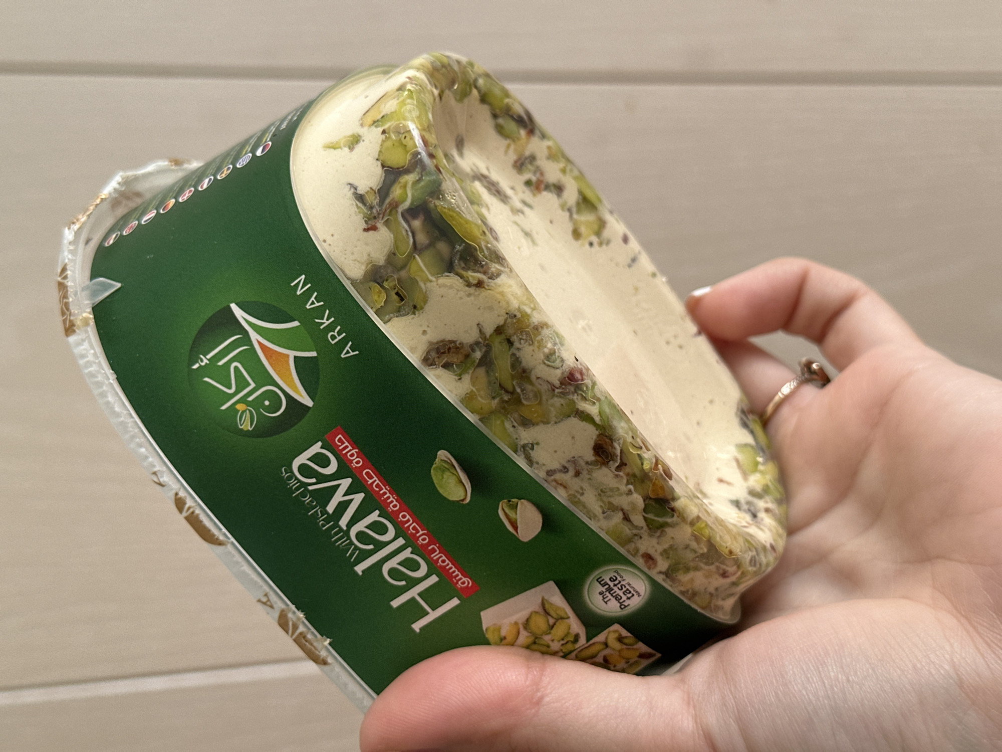The image displays a human hand holding a rectangular plastic container of Arkan Halawa with Pistachios. The container is oriented with its top and label facing the viewer, slightly tilted upwards. The label is predominantly dark green, featuring the brand name "ARKAN" in both Latin and Arabic script, and "Halawa With Pistachios" in Latin script, with "حلاوة طحينية فاخرة بالفستق" (Premium Tahini Halawa with Pistachios) in Arabic. Illustrations of halawa blocks with pistachios and individual pistachios are visible on the label, alongside a circular logo stating "The Premium Taste Natural Food."
The transparent bottom section of the container reveals the halawa product, a pale, creamy matrix densely interspersed with numerous green pistachio pieces. The top edge of the container shows a partially detached or torn metallic-colored seal.
The hand, with light skin tone, holds the container with its fingers and palm on the left and the lower part of the hand and ring finger on the right. A ring with a textured, possibly metallic or stone setting, is visible on the ring finger. The fingernails are short and appear natural or with light polish.
The background is a plain, light beige or off-white surface, possibly a wall or table, with a faint vertical line visible towards the right side. There is no visual information within the image to determine the specific city of Chaikovsky, Russia, or any other precise geographical location.
