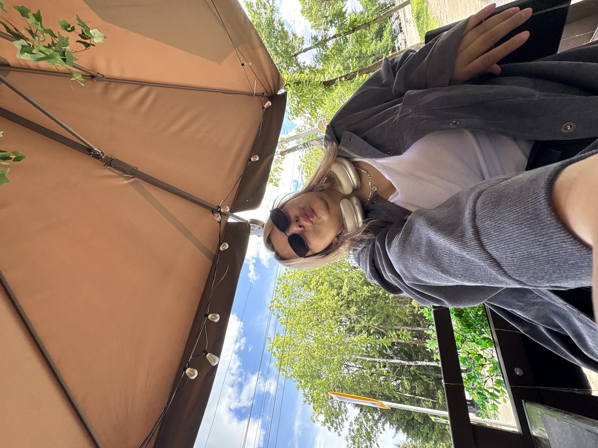 A young woman is sitting outdoors under a large, brown umbrella. She is wearing large, white headphones around her neck, sunglasses, and a grey jacket over a white top. Her lips are puckered as if she is about to kiss. The background shows lush green trees and a bright blue sky with scattered clouds. The scene appears to be from a cafe or outdoor seating area in a city park. No specific landmarks of Birobidzhan, Russia are visible in the image.
