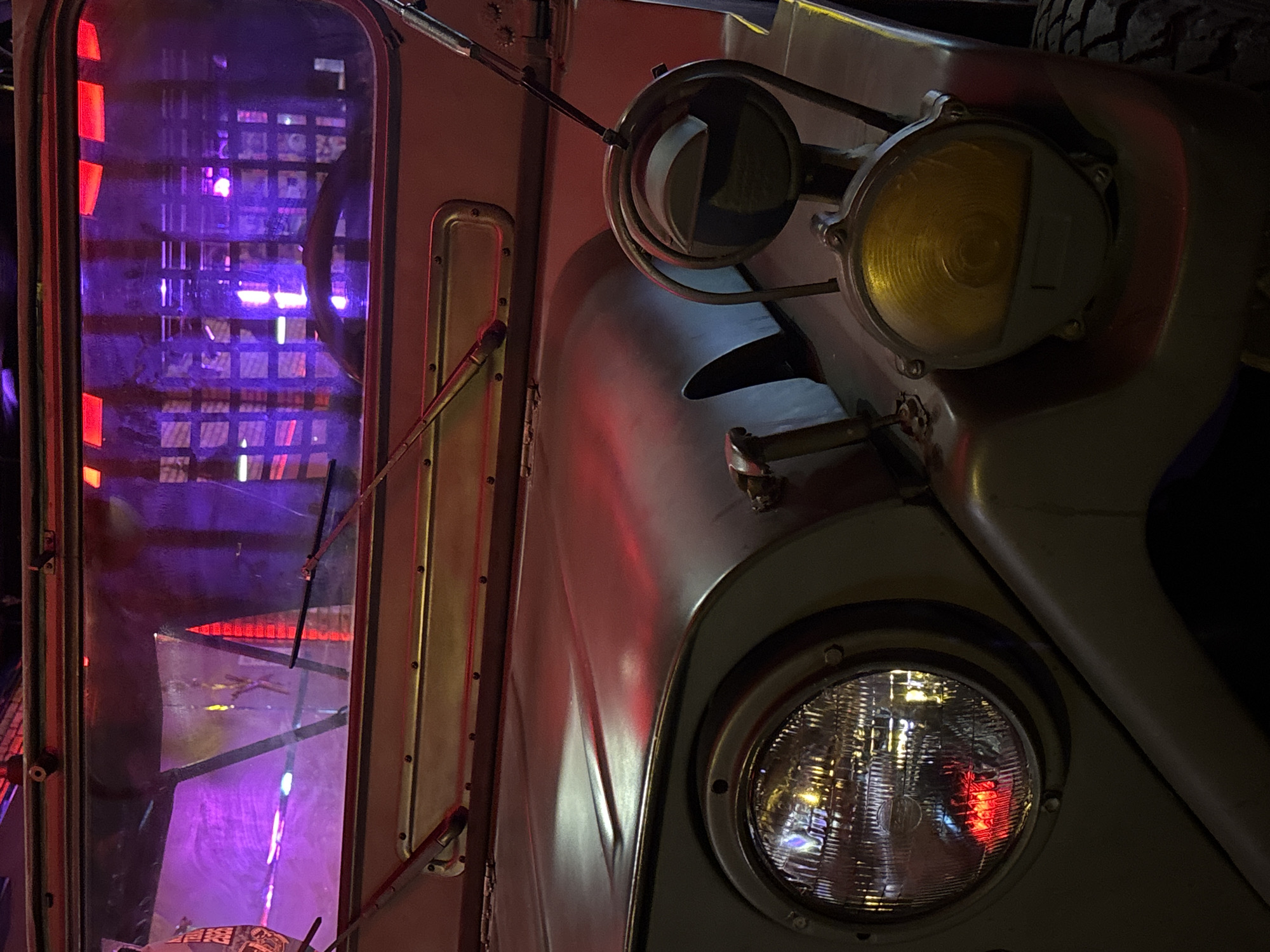The image shows a close-up view of the front of a vintage military-style jeep. The jeep is a muted olive green color. The large, round headlight on the left side of the image is clear and reflects the surrounding lights, including a red light. To the right of the headlight, there is a smaller, circular amber fog light. The hood of the jeep is visible, with a raised central ridge. Above the hood, the windshield wipers are in the down position. The windshield itself is reflecting purple and red lights, suggesting an indoor environment with colored lighting, possibly a bar or an entertainment venue. The background visible through the windshield is a lattice-like structure illuminated with purple and red lights. The overall impression is one of a preserved or displayed vehicle in a dimly lit, atmospheric setting. No people are visible in the image.