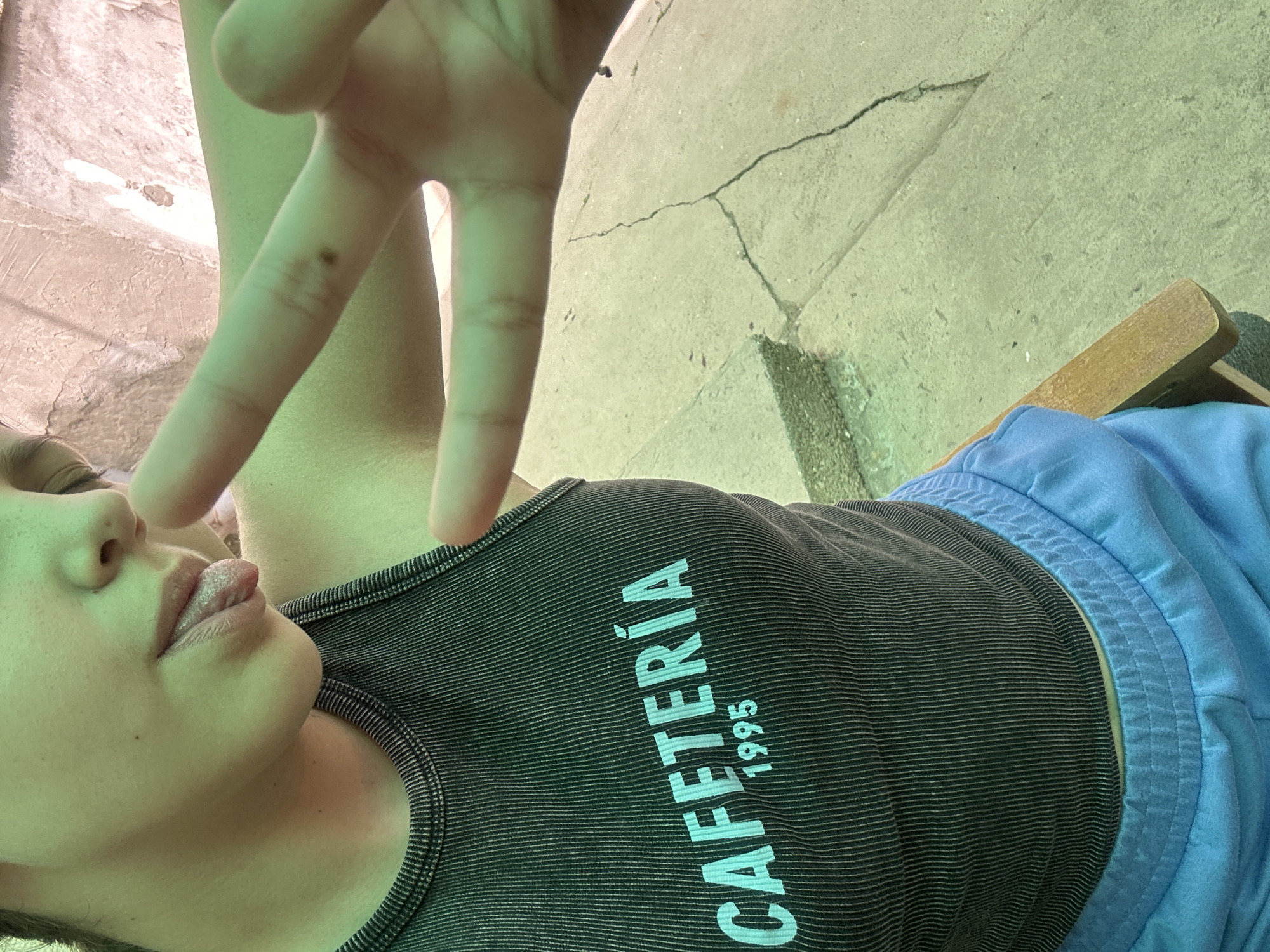 A person, partially visible from the chest up, sits in a wooden chair outdoors, likely in the city of Govedare, Bulgaria. The person is wearing a dark, ribbed tank top with "CAFETERÍA 1995" printed in light blue lettering across the chest. Blue sweatpants are visible below the tank top. The person's left hand is raised, forming a peace sign, and their tongue is slightly extended. The background consists of cracked concrete pavement. The lighting suggests it is daytime, with a green hue cast over the scene.
