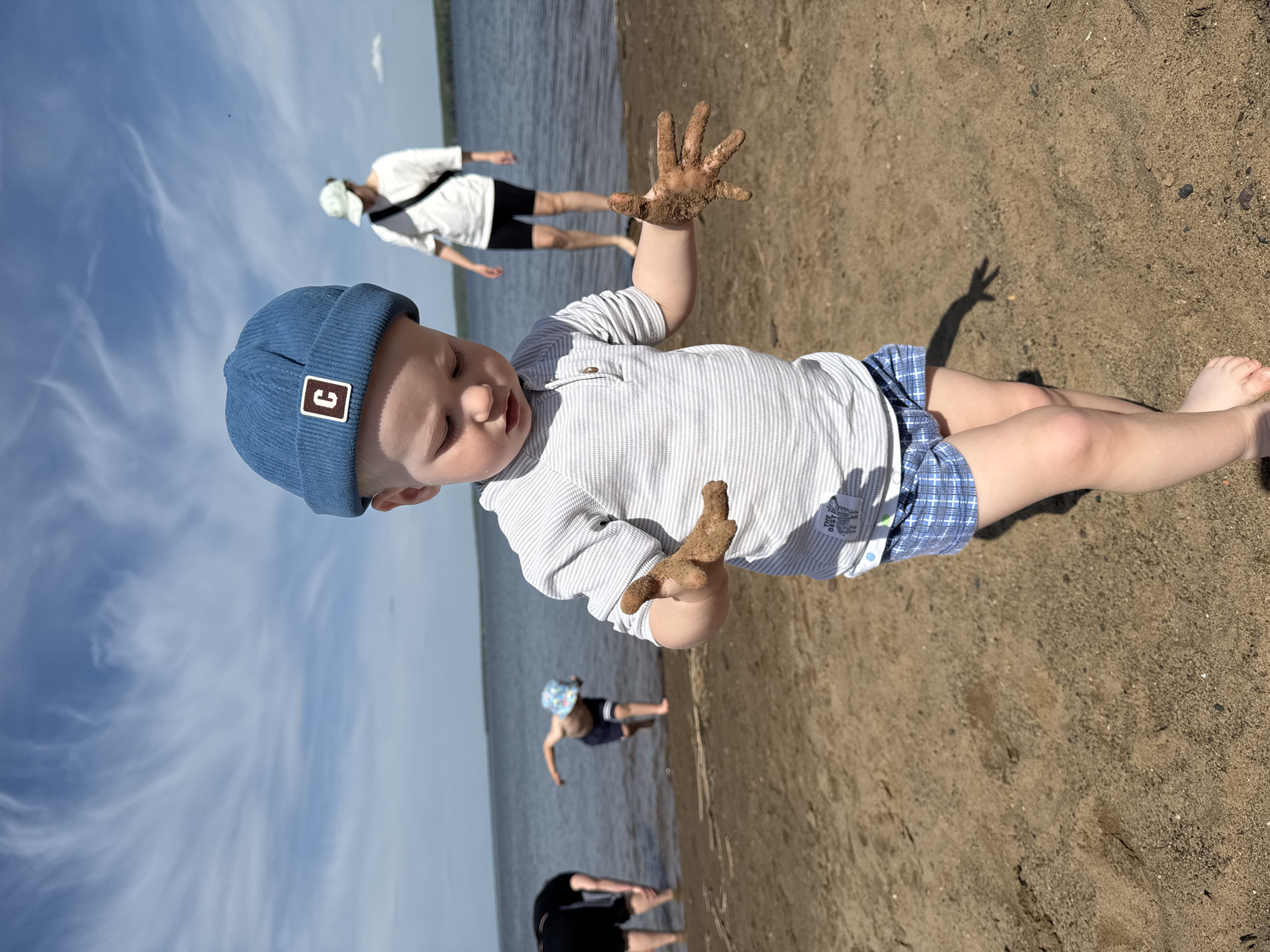 A young child is the central subject of the outdoor photograph. The child, wearing a blue cap with a "9" logo, a striped shirt, and blue plaid shorts, is positioned diagonally on a sandy beach. The child's hands are covered in sand and held in front of them. The legs of two other individuals are partially visible at the bottom of the frame, in the water. Further away, two other people are visible: one in the water wearing a floral cap, and another standing near the water's edge wearing a white shirt and dark shorts. The sky is visible in the background. The setting appears to be a beach on the shore of a body of water, likely a lake or reservoir, in Chaikovsky, Russia.
