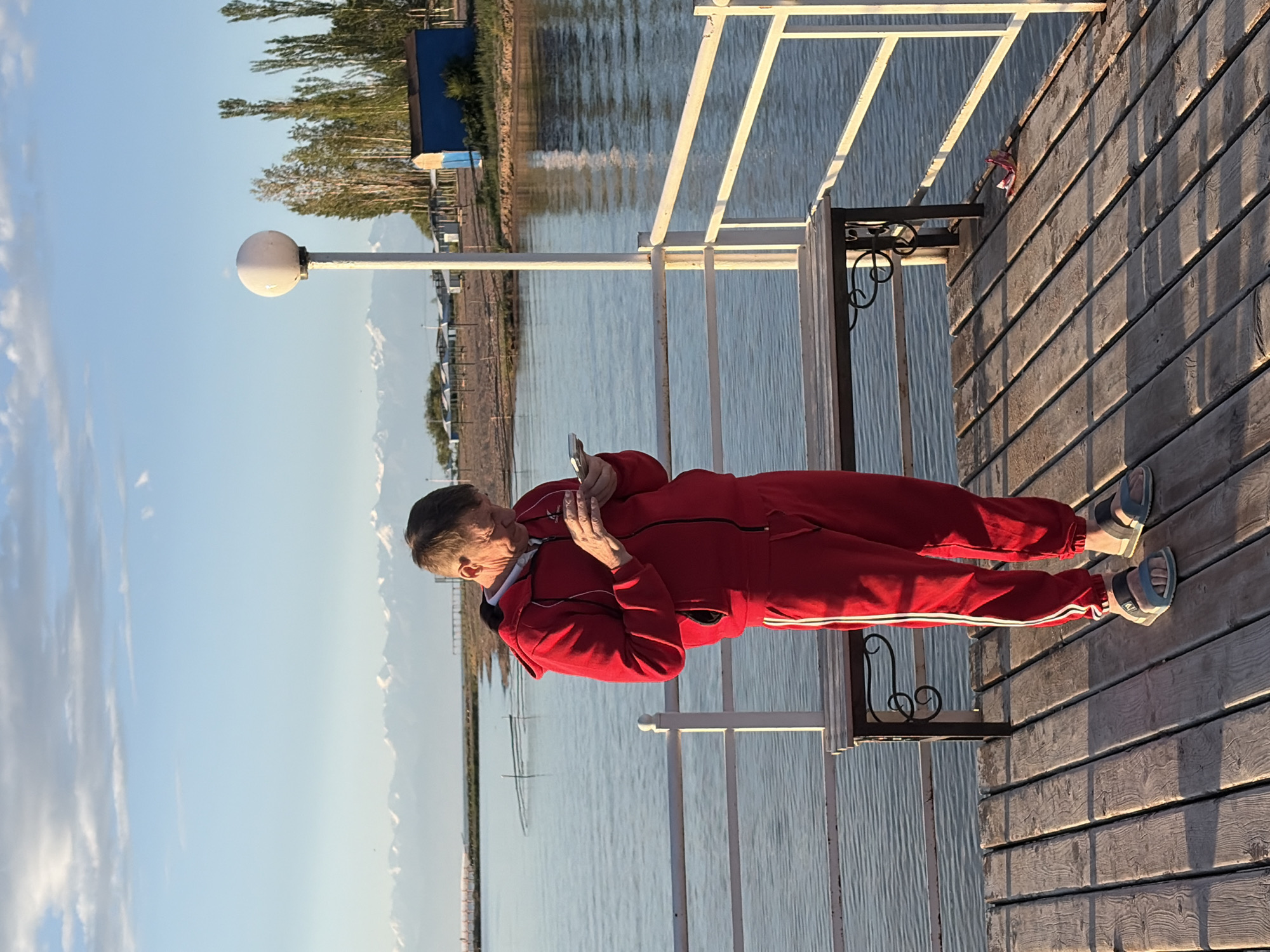 An adult male is standing on a wooden pier, looking at his cell phone. He is wearing a red tracksuit and sandals. The pier is located on a body of water, with mountains in the background. A lamppost is also visible on the pier. The sky is clear and blue, with some clouds. The scene appears to be a recreational area, possibly a park or a resort.