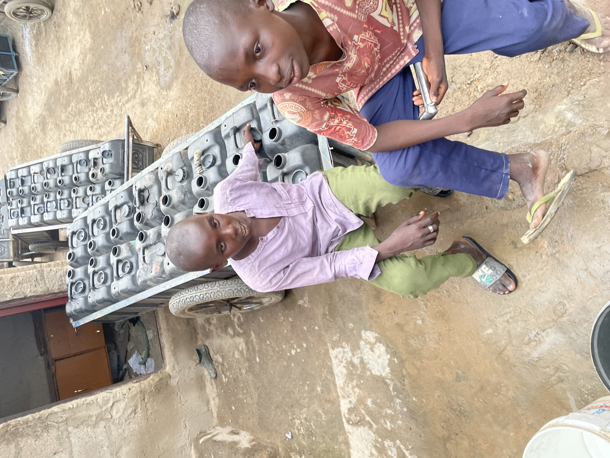 Two young boys are near a cart filled with black containers with openings. One boy, wearing a red and brown patterned shirt and blue pants, sits on the cart and holds a metal object. The other boy, wearing a purple shirt and green pants, lies on the cart. They are in a location with a muddy ground, a building with an open doorway, and other objects scattered around. Location: Unknown, Unknown.