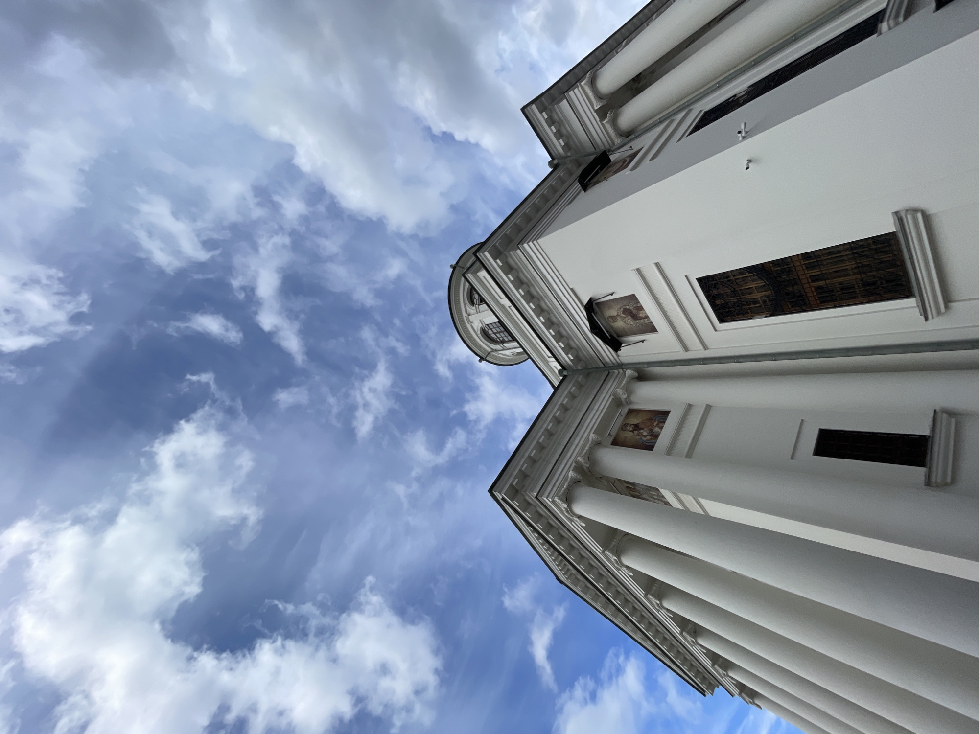 The image captures a low-angle perspective of a grand, white architectural building, likely a cathedral or a historical public structure, set against a bright, partly cloudy sky. The building, viewed from below and to its side, occupies the right and lower portions of the frame, tilting diagonally.
The primary subjects are the impressive architecture and the dynamic sky. The building features numerous tall, white columns, suggesting a classical or neo-classical style. Intricate cornices and moldings adorn the facade and roofline. Several rectangular framed paintings or frescoes, depicting religious figures such as the Madonna and Child, are integrated into the upper walls between sections of the building, adding to its ornate character. Dark, recessed windows, some appearing to have decorative grilles, are also visible. Small security cameras are mounted on the white walls.
The activity depicted is merely the static presence of the building under the sky. There are no people visible, and no events are taking place within the frame.
Notable details include the clear daytime setting, indicated by the bright illumination and distinct cloud formations. The sky is a vibrant blue, filled with a mix of fluffy white cumulus clouds and thinner, wispy clouds, suggesting fair weather. The dramatic low-angle shot emphasizes the height and grandeur of the structure, common in historical and religious buildings, aligning with the location context of Kazan, Russia. The composition creates a sense of scale and awe. No discernible text is visible on the building or in the sky.