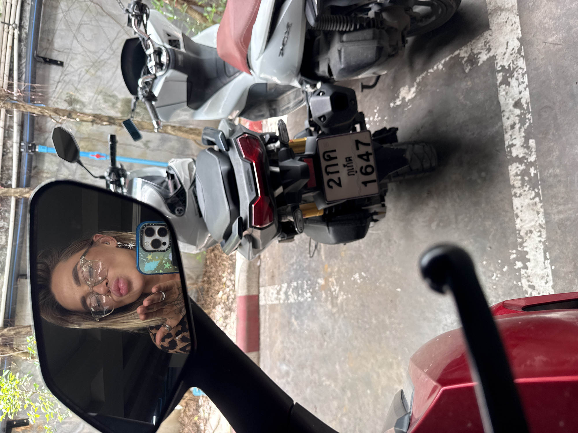 In Choeng Thale, Thailand, a woman is captured in a side-view mirror of a red motorcycle, blowing a kiss. She wears glasses and a leopard-print top. Her left hand, adorned with rings and tattoos, is extended towards the mirror. A phone with a colorful case is held in her right hand. The background reflects a concrete wall with vines and two white scooters parked on a paved surface marked with white lines. One scooter displays a license plate with Thai characters and the number "1647".