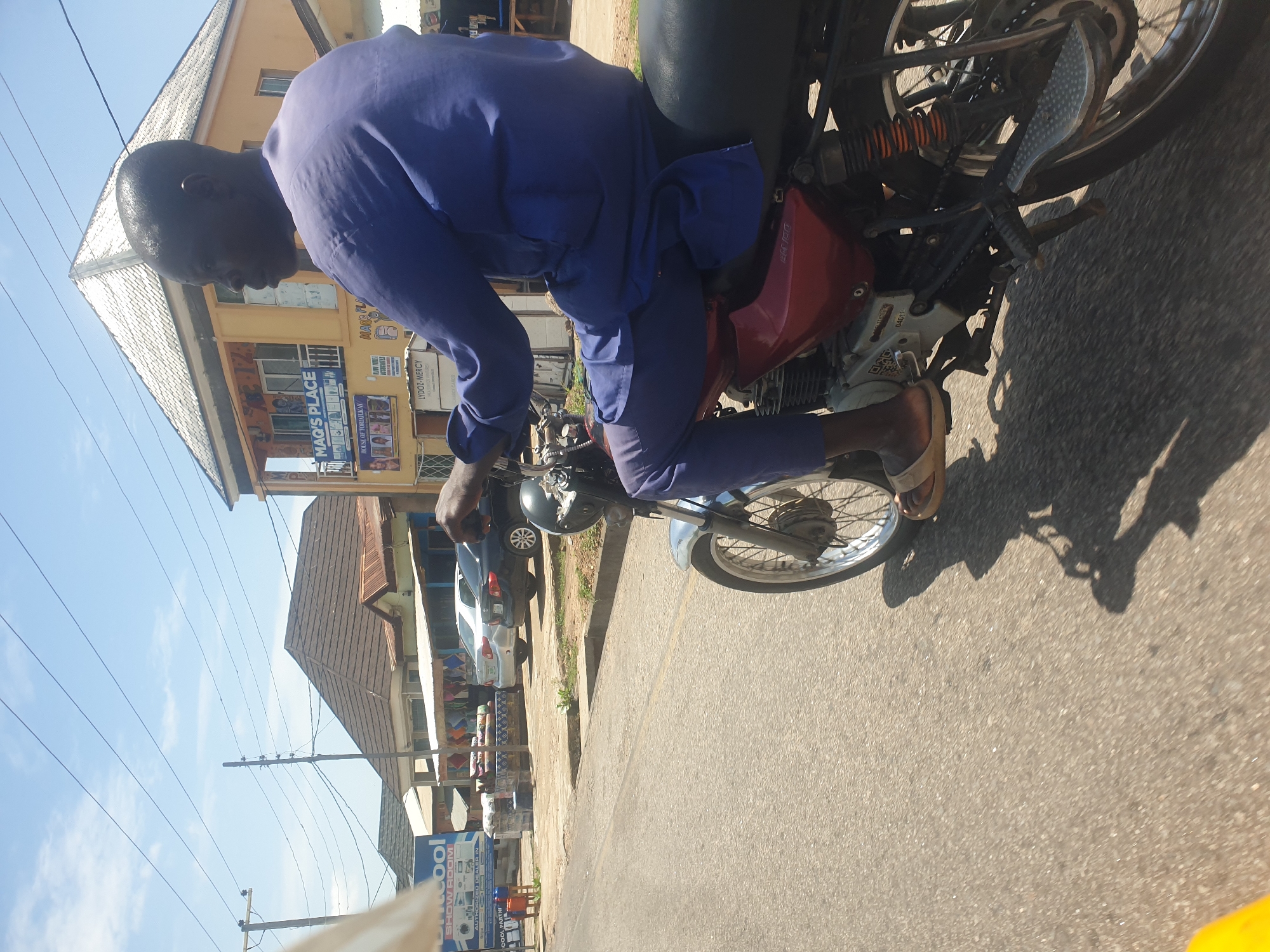 A man is riding a motorcycle in the city of Unknown, Unknown. He is wearing a blue shirt and pants and sandals. The motorcycle is red and black. The road is paved and there are buildings on either side. There is a Unicool showroom, Mag's Place, and Mar's Place.