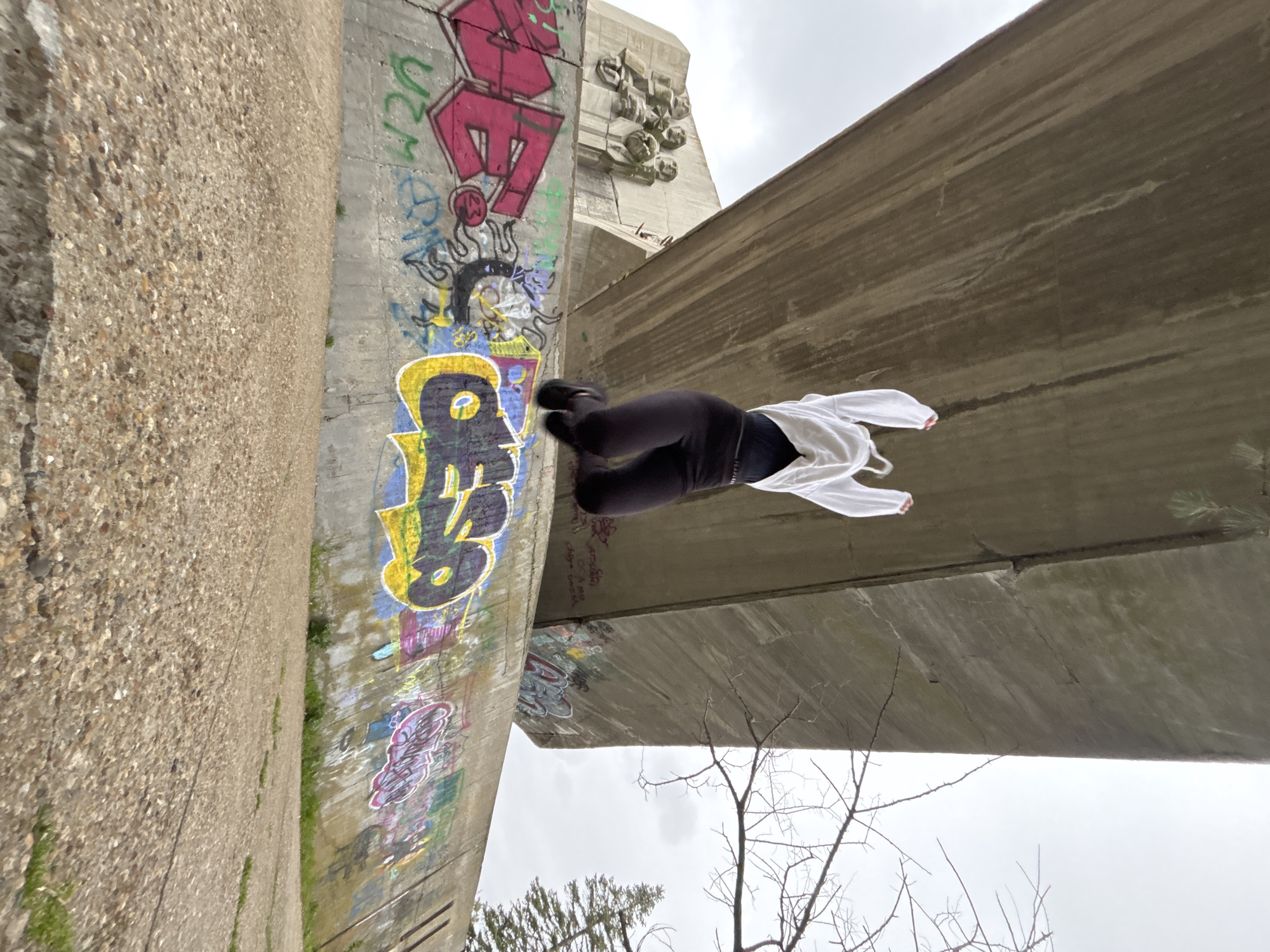 A low-angle view of a person in black leggings and a white hoodie perched on a concrete structure with graffiti. The graffiti is on the left and features colorful designs. The concrete structure appears to be part of a bridge or overpass. The person is positioned horizontally, giving the impression of defying gravity. Bare tree branches are visible in the lower right against a cloudy sky. The location is tagged as Unknown, Unknown.