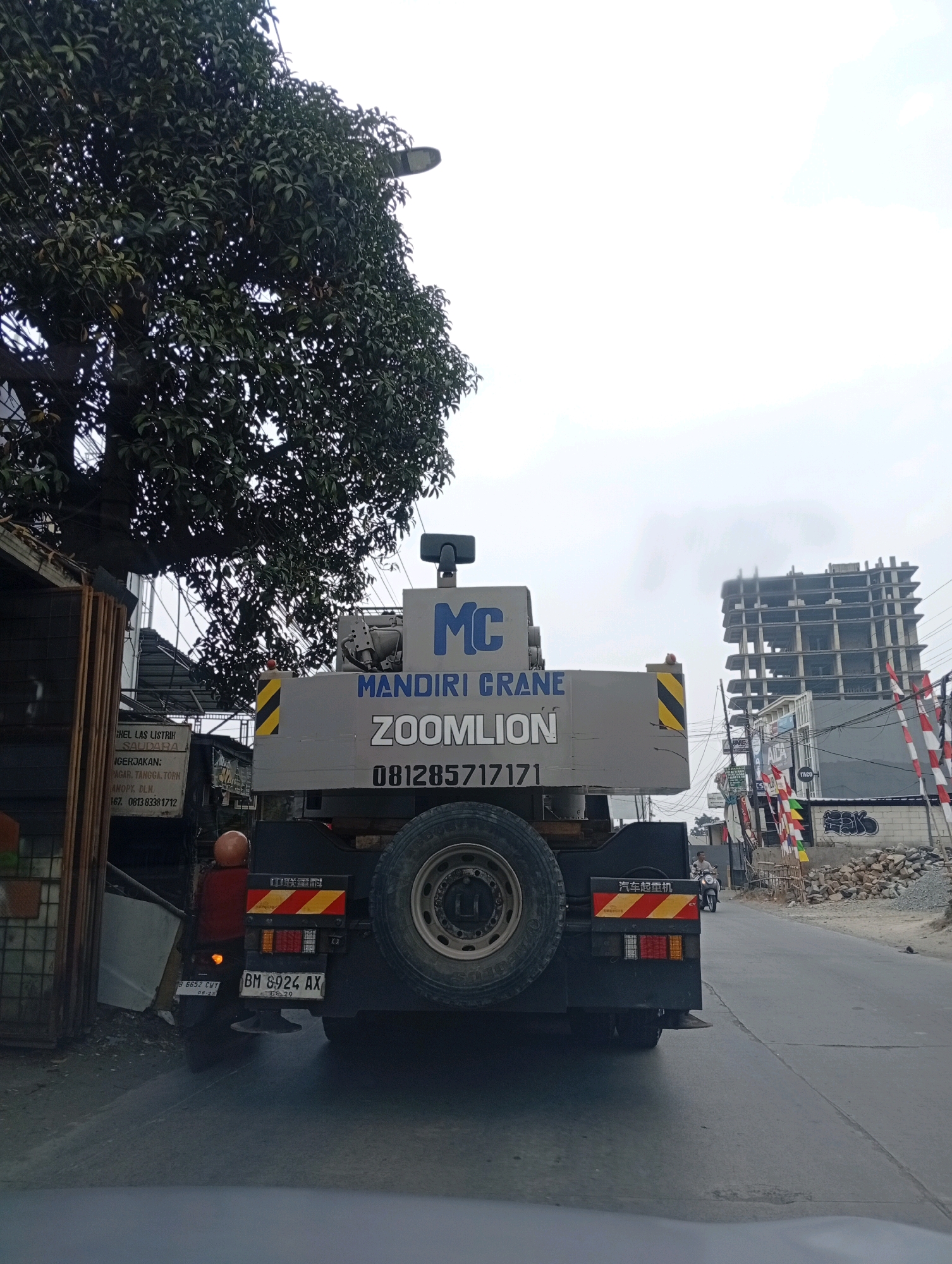 A truck with a crane is parked on a street in Kelapa Dua, Indonesia. The truck is gray and has the words "MANDIRI CRANE ZOOMLLION" written on the side. The license plate of the truck is "BM 8924 AX". There is a partially constructed building in the background, and several red and white flags are flying. A tree with dark green leaves is on the left side of the image. A person on a motorcycle is riding past the truck. The sky is overcast.