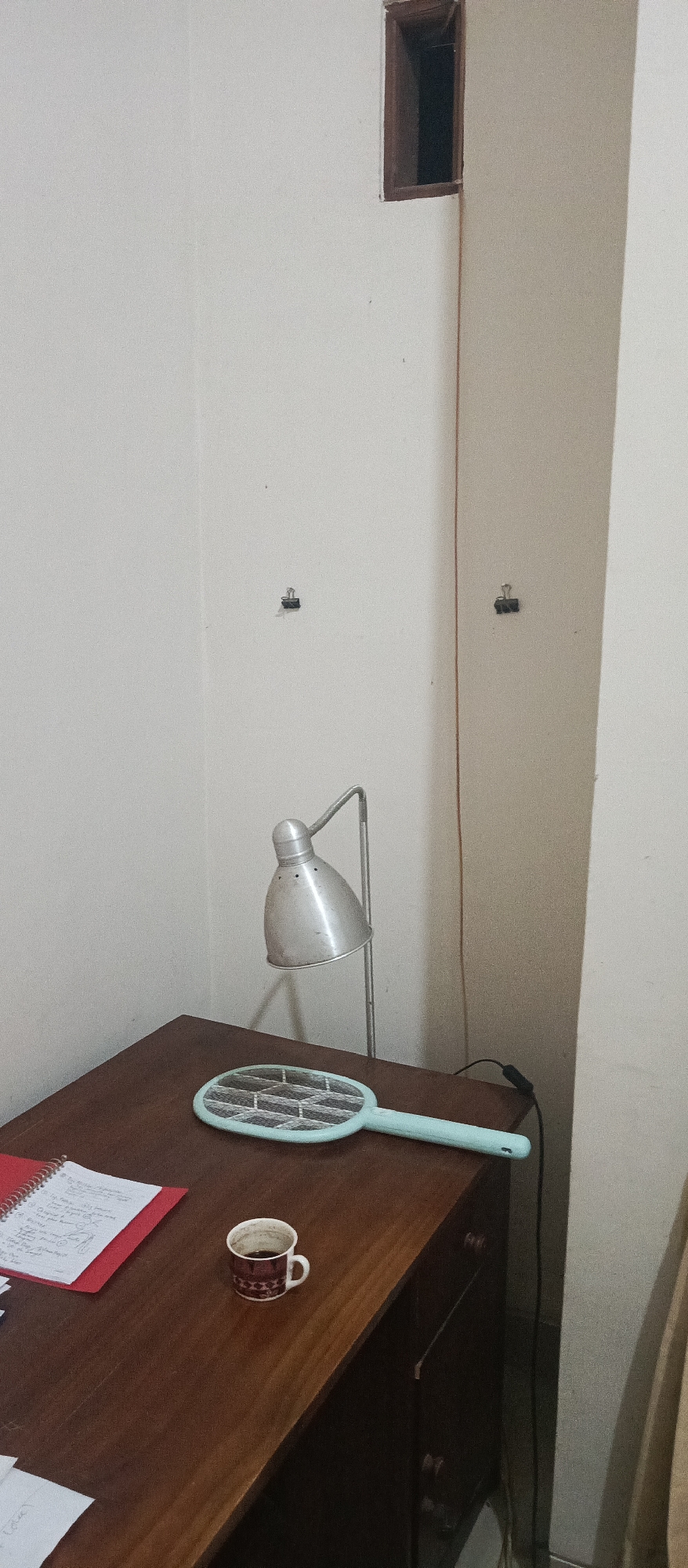 An indoor, eye-level, full shot captures a corner of a room. The walls are painted a light beige. On the right wall, near the top, is a dark, recessed opening, possibly a small window or vent, framed by dark wood. A thin, dark cord runs vertically down the wall from this opening. Two black binder clips are attached to the wall, one higher than the other and spaced horizontally.
A wooden desk occupies the lower portion of the frame. On the desk sits a silver desk lamp with an adjustable gooseneck and a conical shade. Next to the lamp is a light blue electric mosquito swatter with a hexagonal grid. To the left of the swatter is a white coffee mug with a brown and white patterned rim, filled with dark liquid. A red notebook with spiral binding lies to the left of the mug, open to a page with handwritten notes. A corner of a white sheet of paper is visible in the bottom left. The desk appears to have drawers on the right side, one of which is slightly ajar. The lighting in the scene is dim, with a shadow cast by the lamp on the desk.