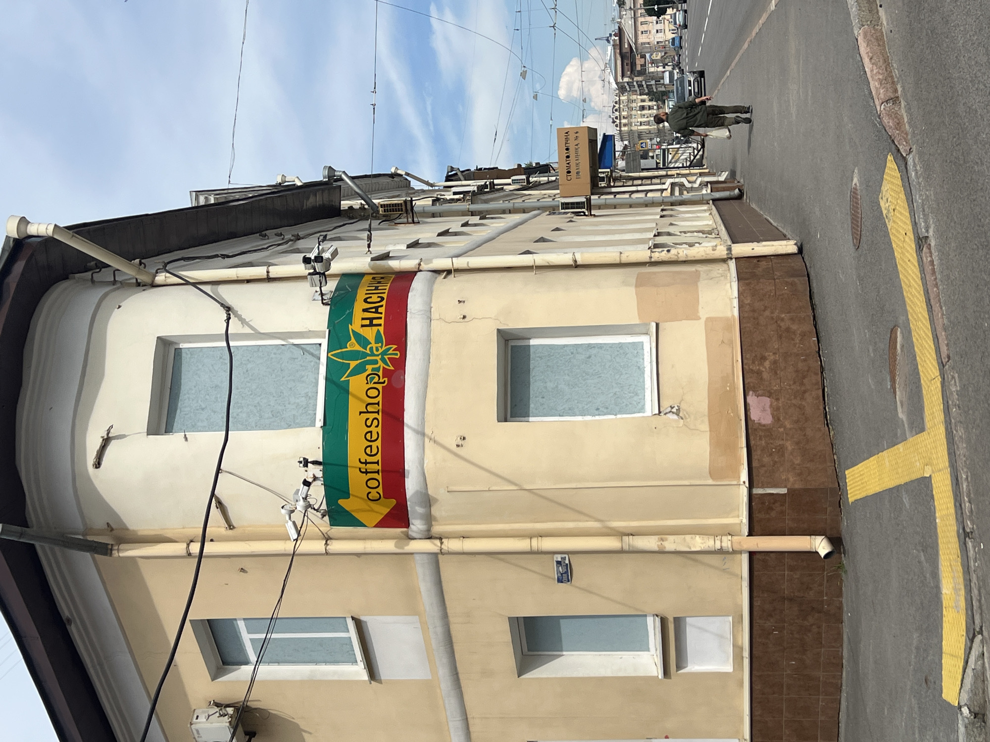 The image shows a corner building on a city street in Kharkiv, Ukraine. The building is painted a pale yellow with a brown tiled base. A sign with a yellow arrow pointing left reads "coffeeshop" in red and green lettering, with a cannabis leaf graphic above it. The building has several windows, some covered with opaque blue material. Security cameras are mounted on the building. Further down the street, a person in a dark green uniform is walking away from the camera, carrying a white plastic bag. The street appears to be a multi-lane road with traffic. There are overhead wires and streetlights visible. The sky is partly cloudy.