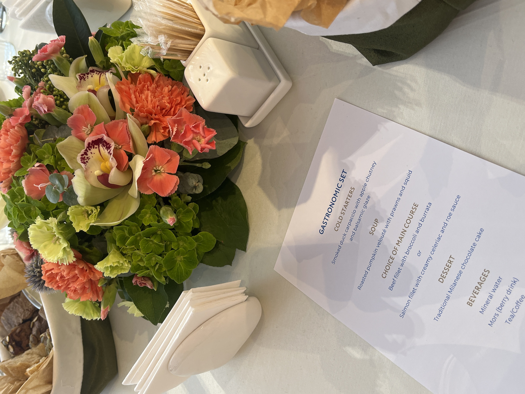 The image displays a close-up, elevated view of a formal or semi-formal dining table setting, likely at an event in Shushary, Russia. The scene features a vibrant floral centerpiece, a menu, and various table essentials.
The floral arrangement is rich with color and texture, comprising prominent light green and pink orchids with dark red accents, numerous ruffled orange and coral carnations, and abundant green foliage, including small green berry clusters.
Around the centerpiece, a minimalist white salt shaker rests on a rectangular tray, and a stack of white paper napkins is neatly held in a dispenser. Partially visible on the left is a dish containing brown, textured food items, possibly bread or pastries, and in the background, a clear plastic bag holds wooden skewers. A dark green fabric, likely a napkin, is also present. The table is covered with a light-colored tablecloth.
The white menu, titled "GASTRONOMIC SET," offers a multi-course meal:
- **Cold Starters:** Smoked duck carpaccio with apple chutney and balsamic glaze.
- **Soup:** Roasted pumpkin velouté with prawns and squid.
- **Choice of Main Course:** Beef fillet with broccoli and burrata, or Salmon fillet with creamy celeriac and roe sauce.
- **Dessert:** Traditional Milanese chocolate cake.
- **Beverages:** Mineral water, Mors (berry drink), and Tea/Coffee.
The inclusion of "Mors (berry drink)" on the menu aligns with the provided location context of Russia. The overall setting suggests a pleasant and curated dining experience.