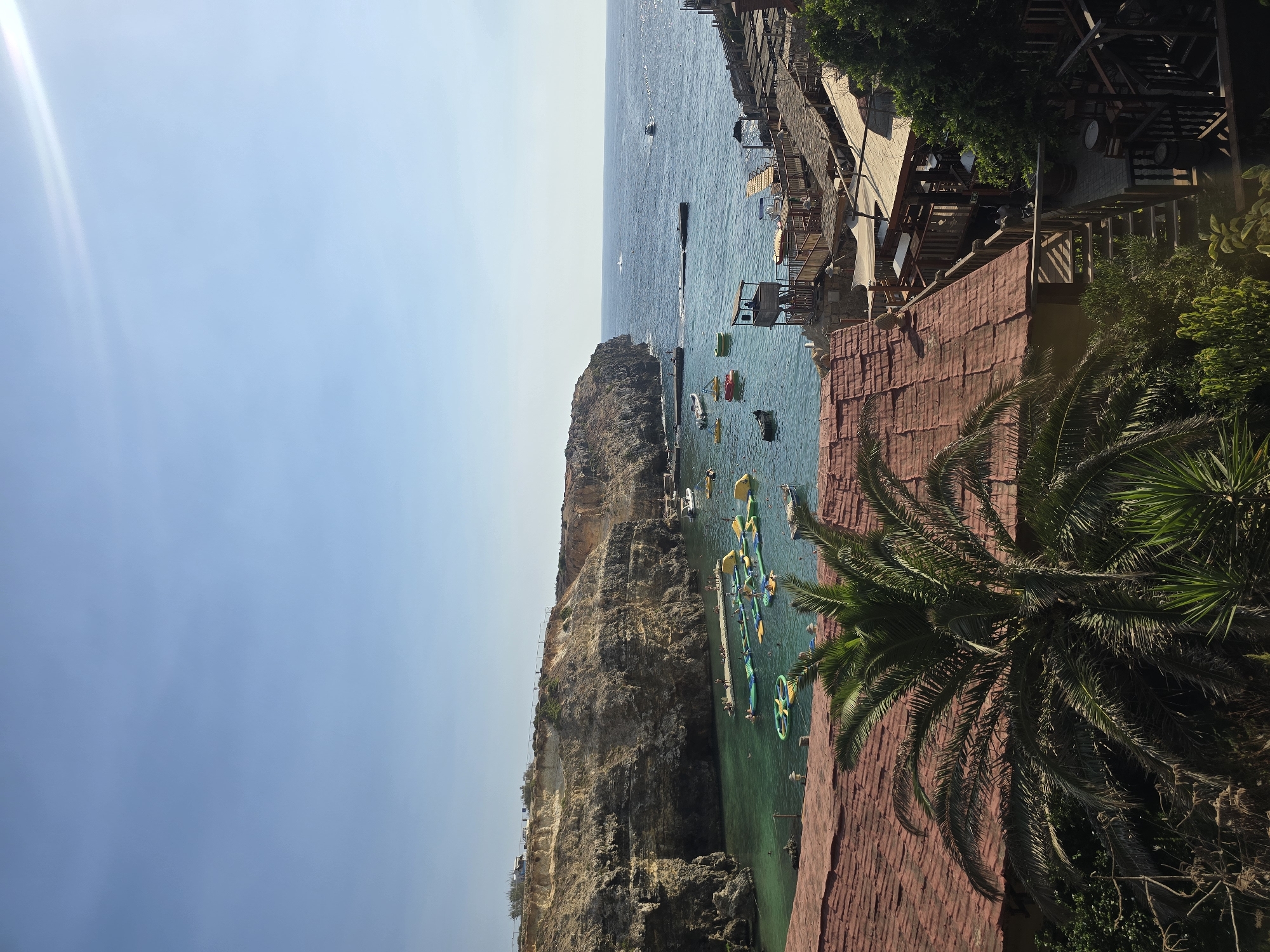 This image depicts a scenic view of a coastal area, likely a bay or inlet, in Mellieħa, Malta. The scene is bathed in clear sunlight under a pale blue sky. In the foreground, the reddish-tiled roofs of several structures are visible, partially obscured by the fronds of a large palm tree.
The midground features a vibrant turquoise bay, dotted with various inflatable water attractions, including slides and climbing structures, suggesting a recreational area. Several small boats are moored or moving within the bay. People can be seen on a pier extending into the water, interacting with the inflatable structures, and swimming. Cliffs rise steeply on the left side of the bay, showcasing layered rock formations and sparse vegetation.
On the right side of the bay, a series of wooden decks, stairs, and thatched-roof structures are built into the rocky terrain, indicating a waterfront establishment with seating areas and possibly a restaurant or bar. The background shows the open sea stretching to the horizon, with a few distant boats. The overall impression is of a lively, sunny day at a popular coastal leisure spot.