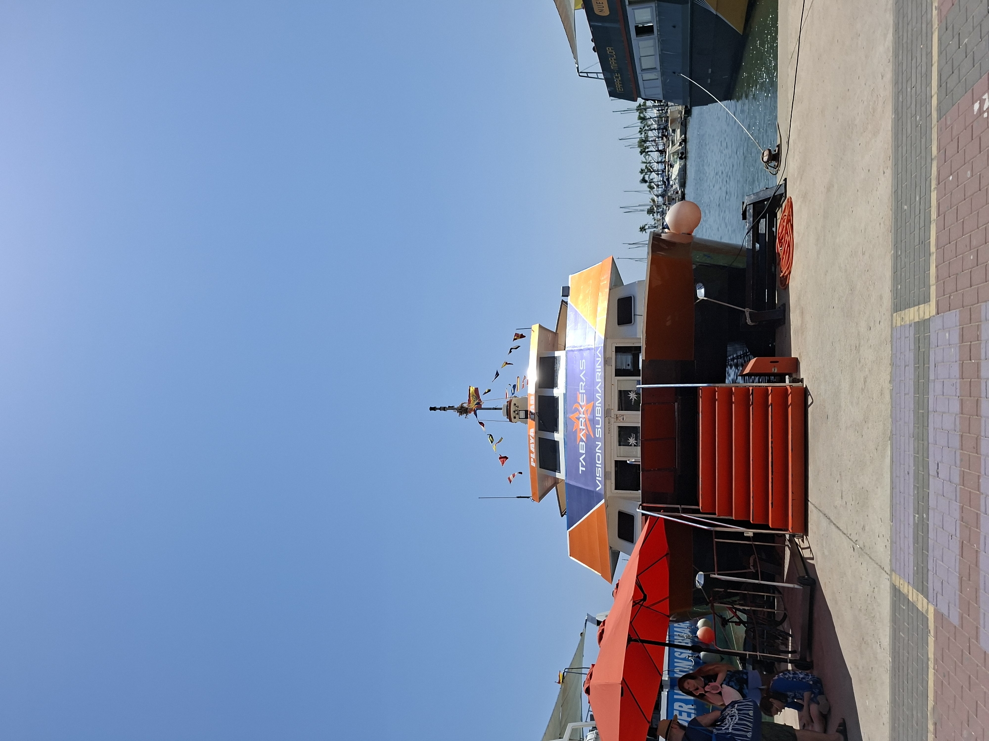 This is a vertically oriented, full-shot image taken in Santa Pola, Spain, likely during daylight. The scene is a waterfront, with a clear blue sky above. In the foreground, a bright orange umbrella is open, casting a shadow on a paved area. To the left of the umbrella, several people are seated or interacting, some with their backs to the camera.
Dominating the mid-ground is a multi-story boat or ferry with an orange and blue color scheme. Prominent text on its side reads "TABARKERAS" and "VISION SUBMARINA", indicating it's likely a tour boat, possibly for underwater viewing. Red steps lead up to the boat's entrance. In the background, several sailboats are docked in a marina, with palm trees visible along the shore. Another larger boat, predominantly dark grey with yellow accents, is moored to the left, with visible text on its hull reading "TERRALE MIRALOR".
The overall impression is a sunny, pleasant day at a harbor, suggesting a tourist area. The presence of people enjoying the outdoors and the docked boats points to a leisure activity or a port in operation.
