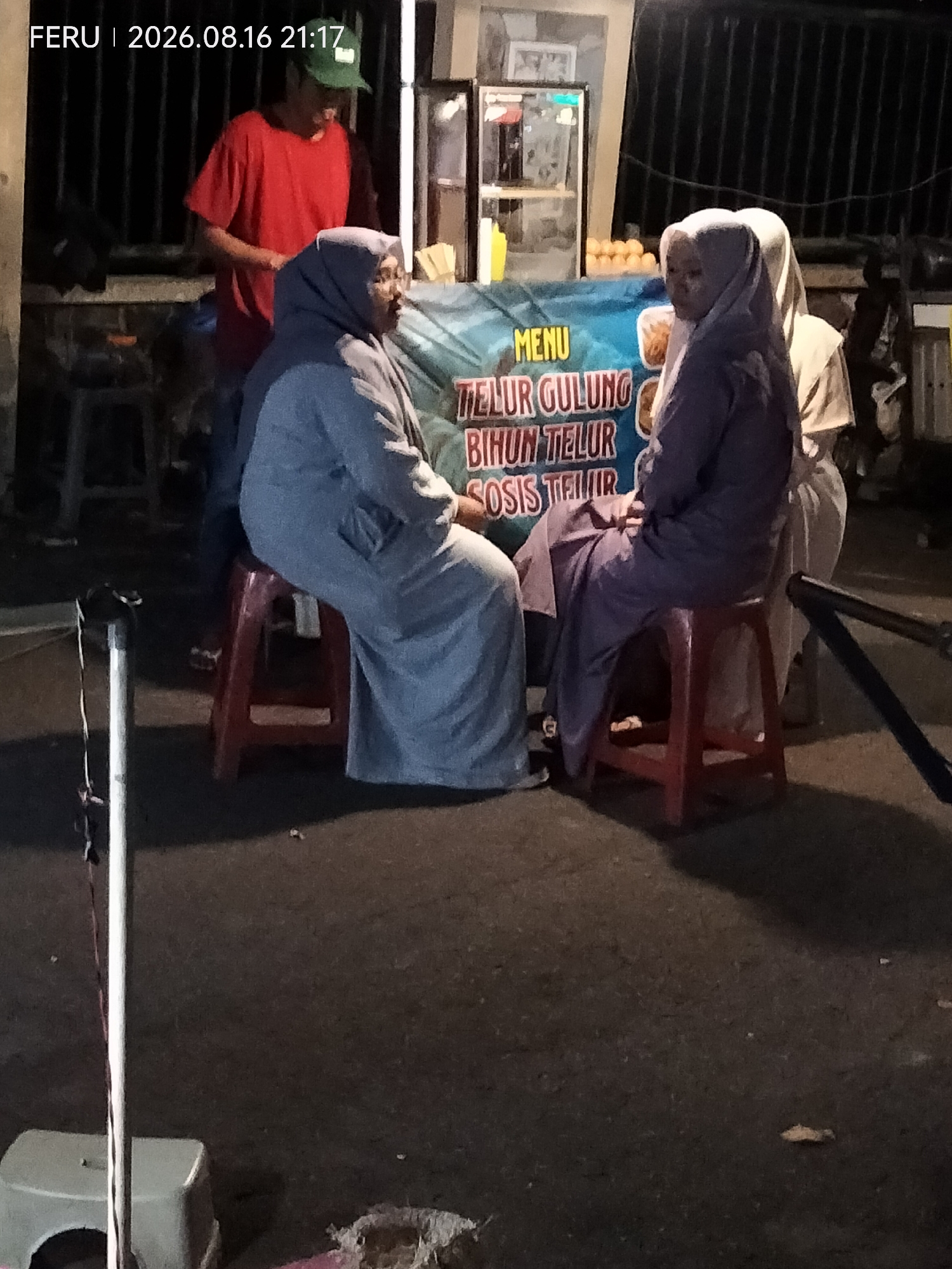 In Rejoagung, Indonesia, a street food vendor stands behind a stall displaying "MENU TELUR GULUNG BIHUN TELUR SOSIS TELUR" in Indonesian. Two individuals, likely customers, are seated on red stools facing the stall, their bodies obscured by their modest clothing and headscarves. One person, on the left, wears a blue headscarf and a long blue garment, while the person on the right wears a purple headscarf and a matching purple garment. The background is dimly lit, with a glimpse of a glass-fronted display case to the left, suggesting a small food stand operating at night. The ground is paved and appears to be a street or sidewalk.