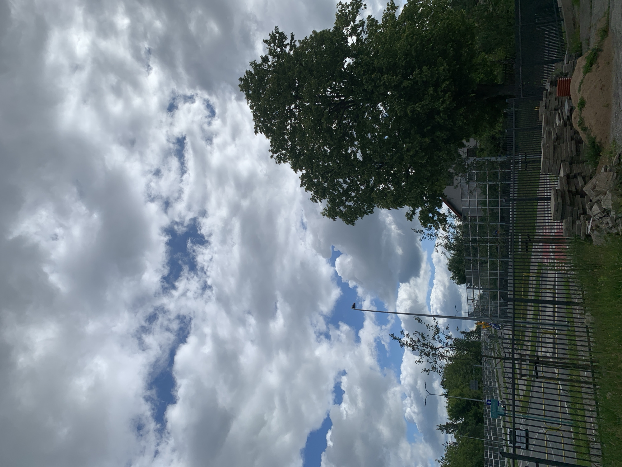 The image shows a cloudy sky with patches of blue. In the foreground, there is a black metal fence. Behind the fence, there is a pile of paving stones and sand. Beyond that, there is a tree-lined street with traffic lights and a building in the background. A white car is visible on the street. The scene appears to be a construction site or an area undergoing development. The location is likely in the city of Białystok, Poland, given the presence of Polish street signs and the architectural style of the building.