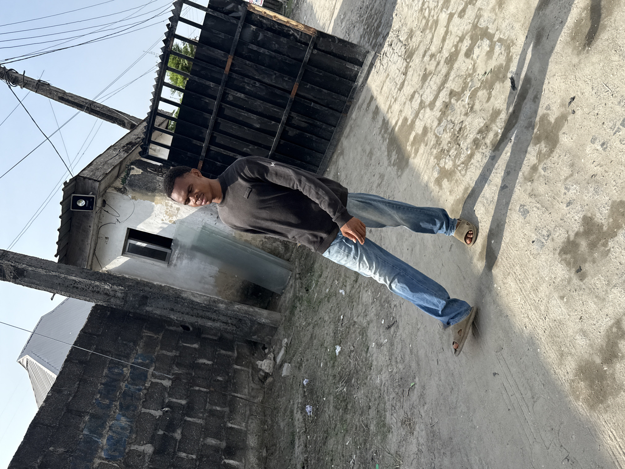 A medium shot of a person in jeans and a black sweater standing on a concrete road is depicted in the photograph. The person is wearing sandals. The setting appears to be urban, with a building featuring a window and a gate nearby. The location is Ado, Nigeria. Power lines are also visible. The image is rotated.