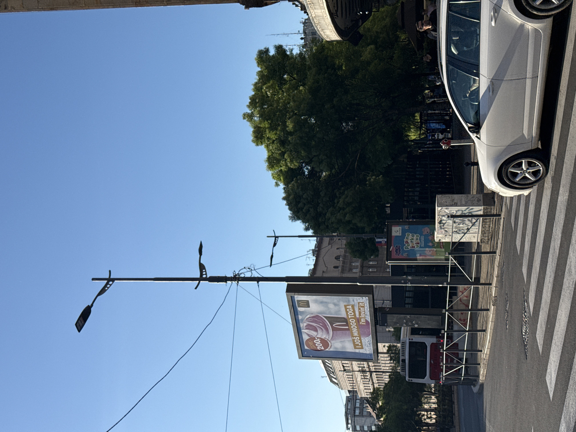 A street in Belgrade, Serbia. A white car is parked on a zebra crossing. Along the street are billboards advertising McDonald's and a Peppa Pig event. Street lights line the road. There are trees and buildings in the background.