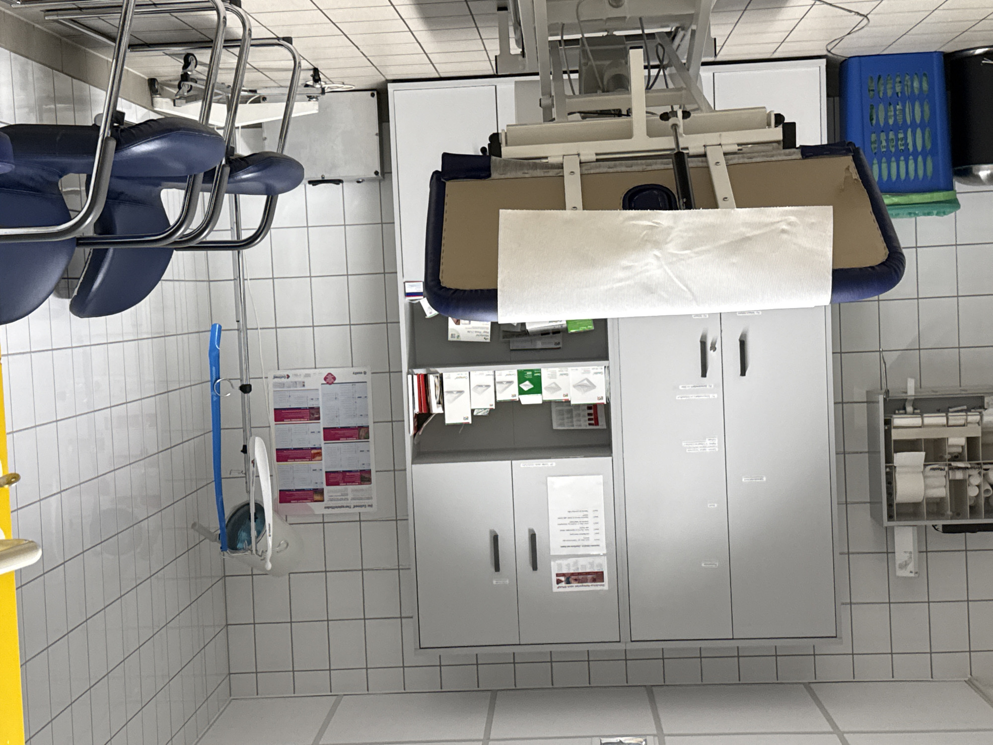 The image depicts an interior scene of what appears to be a medical examination or treatment room. The room is tiled with white square tiles on the walls and floor. In the foreground, a medical examination table with a blue upholstered base and a white paper covering is centrally positioned. To the left of the examination table, a blue plastic laundry basket and a black trash receptacle are visible. Behind the examination table, a large, light grey cabinet with two doors is mounted on the wall. Above the cabinet, a white shelf holds various medical supplies and boxes. To the right of the cabinet, another section of the wall is covered with white tiles. A medical stand with a blue bag and various tubes is also present, along with two dark blue chairs with metal frames. Several posters with medical information are displayed on the wall. The environment suggests a clinical setting, possibly a doctor's office or a clinic, located in the town of Walkenried, Germany.