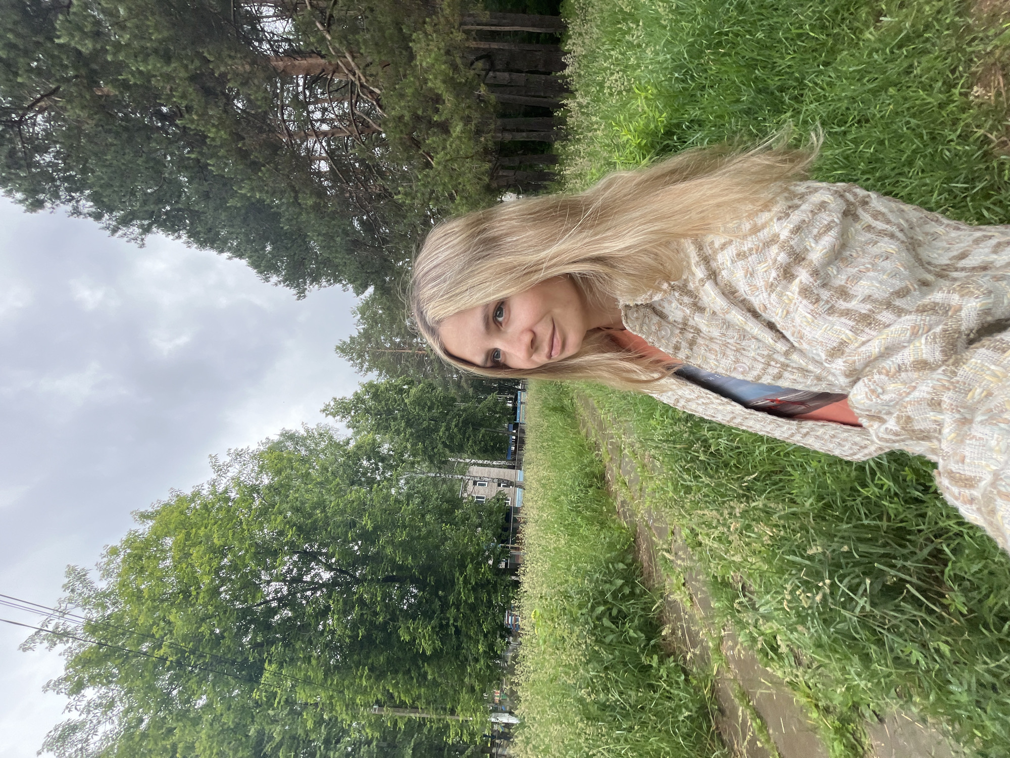 A young woman with long blonde hair smiles gently, looking towards the camera in an outdoor setting. She is wearing a light-colored, textured, possibly woven jacket or cardigan over a darker top, and part of her arm appears to be holding a book or tablet with the partial word "THE" visible on its spine. Her hair is swept to the side, indicating a slight breeze.
The scene unfolds under an overcast sky, suggesting a cloudy day. In the background, tall evergreen trees, likely pines, line the horizon, interspersed with some deciduous trees with lush green foliage. A dirt path or road runs alongside a vibrant green grassy area, which is partially overgrown. Further in the distance, glimpses of buildings and power lines can be seen, indicating a semi-urban or park-like environment rather than a completely wild forest. The overall impression is one of a calm, leisurely moment outdoors on what appears to be a mild, cloudy day in Chaikovsky, Russia.