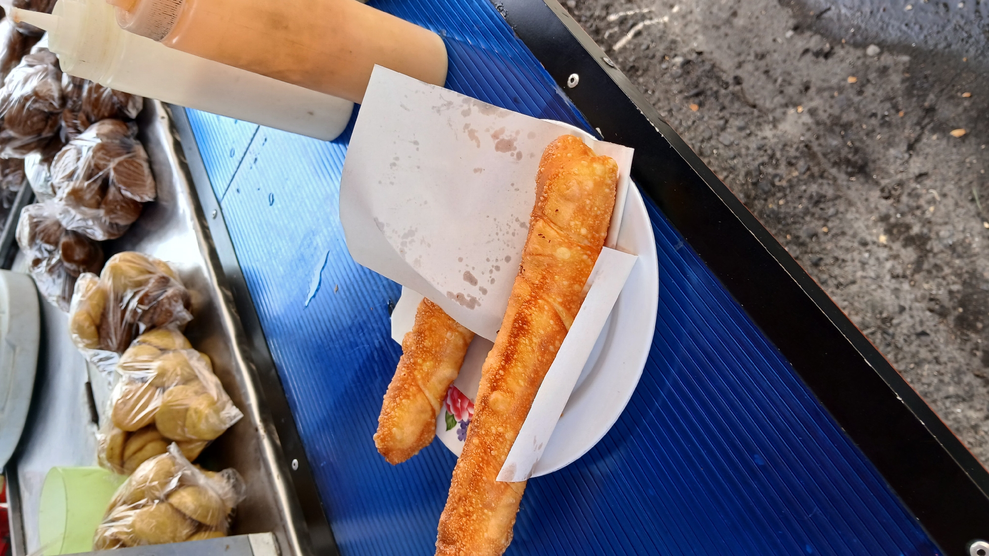 The image shows a food stand in what appears to be Puerto La Cruz, Venezuela. On the stand are packaged pastries, squeeze bottles of sauces, and two fried rolls on a plate with a paper wrapper. The stand itself has a blue corrugated surface with a dark rim. The background shows pavement.