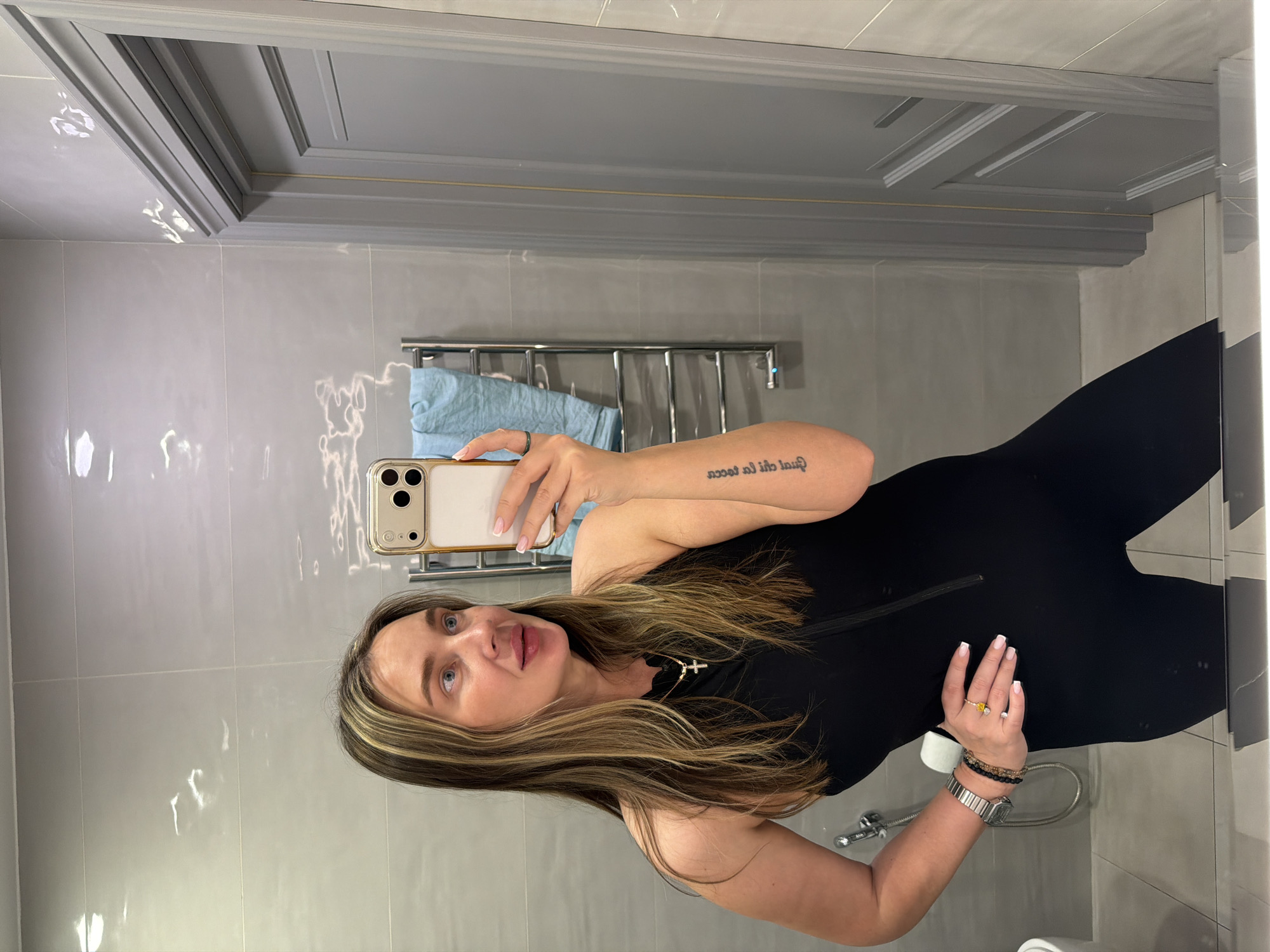A fair-skinned woman is taking a selfie in what appears to be a bathroom. She is wearing a black garment and jewelry on her hands. The bathroom has light gray tile walls and a matching decorative ceiling border. A chrome towel rack with a blue towel hangs on the wall behind her. The location is unknown and cannot be inferred from this image.