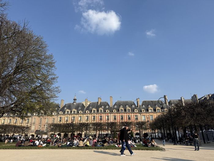 The image captures a daytime scene at Place des Vosges in Paris, France, under a clear blue sky with scattered white cumulus clouds. In the background, the iconic symmetrical architecture of the square is visible, featuring a long row of red brick buildings with stone dressings, numerous windows, and dark grey mansard roofs punctuated by dormer windows and chimneys. Arcades are present at the base of these buildings.
In the middle ground, a large grassy lawn is populated by an estimated 50 to 100 individuals. Most are seated or reclining in small groups or individually, dressed in casual attire, appearing to relax and socialize. A large deciduous tree with visible budding leaves frames the left side of the scene, while other trees, largely bare, line the perimeter of the grassy area. A decorative black street lamp with a spiraling fixture stands prominently near the right side of the lawn.
In the foreground, a wide, light-colored gravel or dirt path extends across the width of the frame. A male subject wearing a dark jacket and blue jeans walks from left to right on this path, carrying a blue object, possibly a folded umbrella or bag. Another individual walks partially obscured behind him. Further to the right on the path, a subject in dark clothing stands with their back to the viewer. Metal barriers are partially visible on the far right edge of the frame.